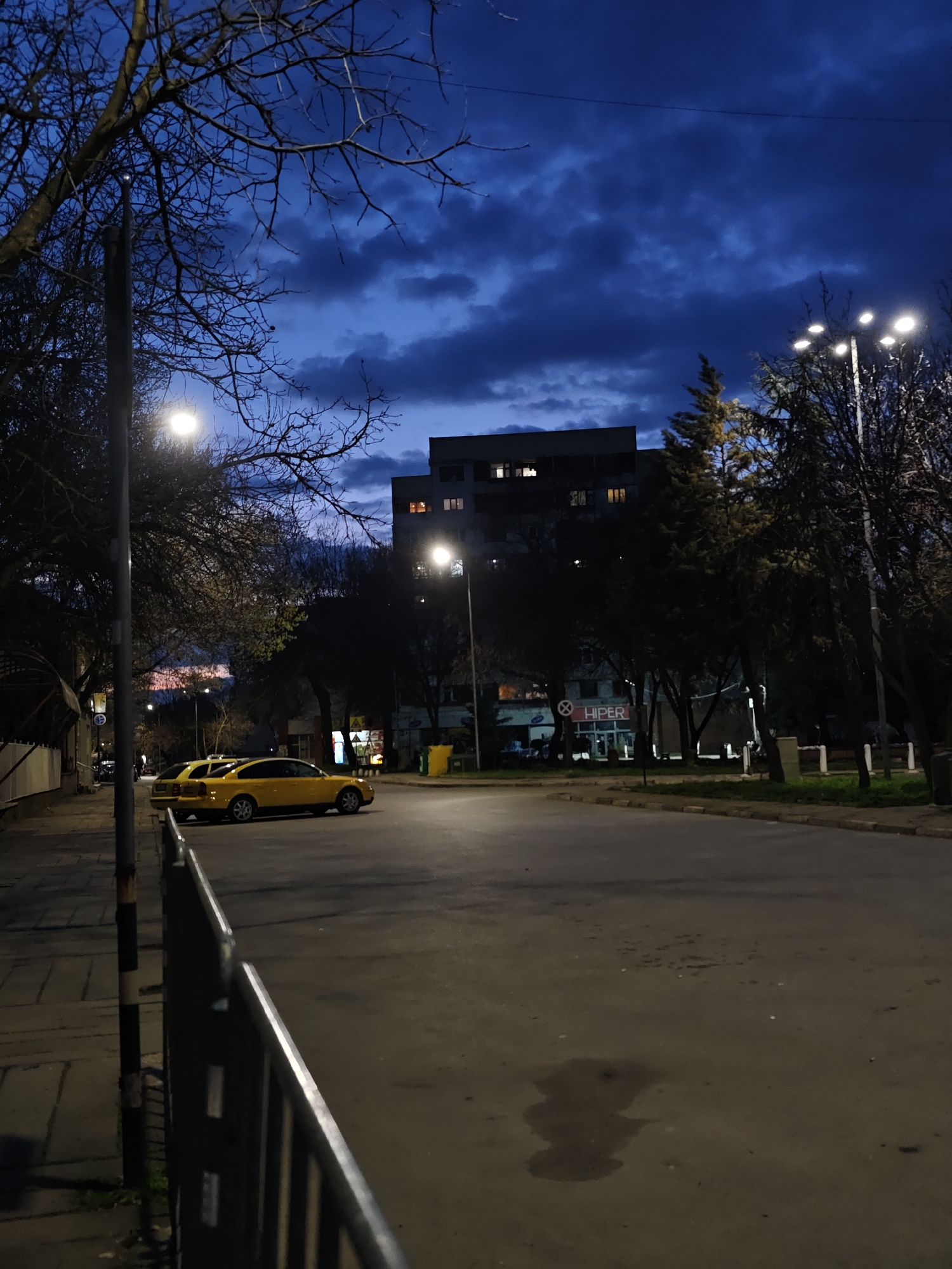 The image depicts a street scene at dusk in what appears to be Kozloduy, Bulgaria. Two yellow cars are parked along the side of the road, illuminated by streetlights. A building with lit windows is visible in the background, as is a store with a sign that reads "HIPER." Trees line the street, some bare and others with foliage. The sky is a dark blue with clouds. A metal fence is in the foreground. The road surface is asphalt with patches of discoloration. No people are clearly visible. The scene is generally quiet and appears to be residential or a quiet commercial area.