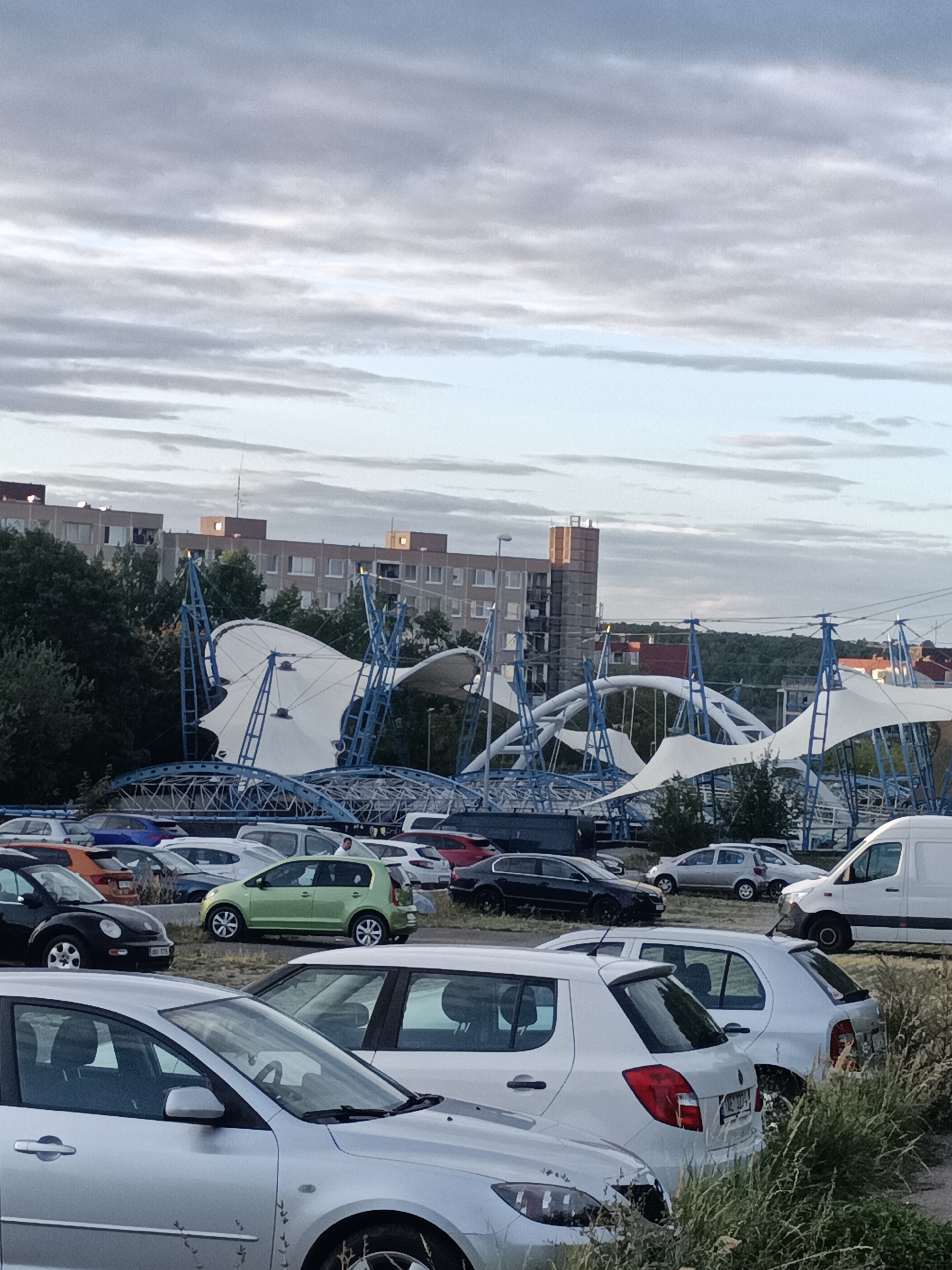 The image depicts a parking lot filled with various cars, set against a backdrop of modern buildings and a distinctive white tent-like structure supported by blue metal frames. The sky is overcast with grey clouds. In the foreground, several cars are visible, including a silver sedan, a white hatchback, and a darker sedan. Behind these, a green hatchback, a black Volkswagen Beetle, and other vehicles are parked. The large, white, curved structure appears to be a temporary event tent or shelter, with its fabric canopy stretched over a complex blue steel framework. Residential buildings, some with balconies and windows, are situated behind trees and the tent structure, suggesting an urban or suburban environment. The overall scene suggests a publicly accessible area, possibly a shopping center parking lot or a venue with outdoor facilities. The specific location in Prague, Czechia, cannot be determined from the visual information alone.