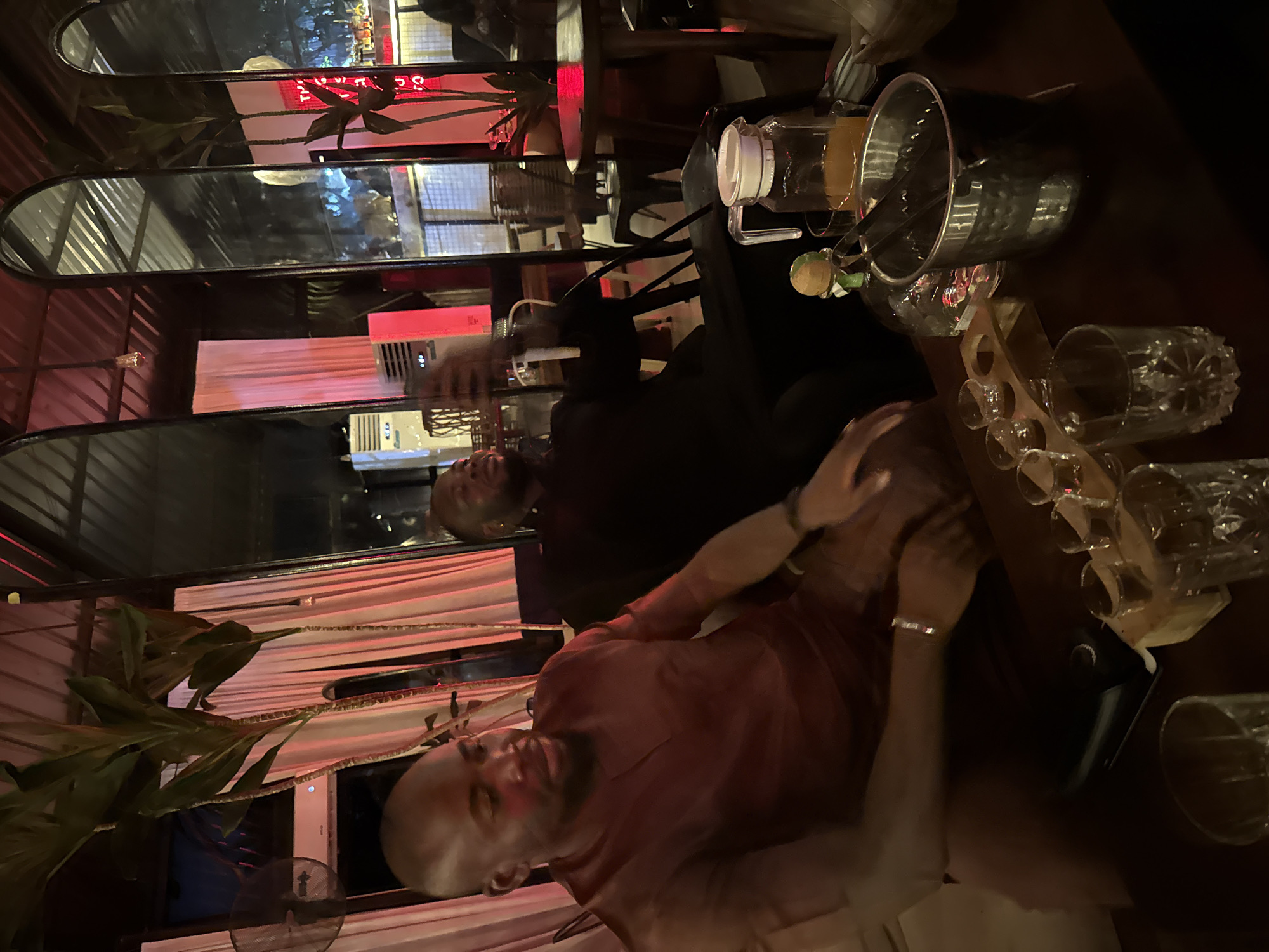 The image captures an interior scene, likely a bar or lounge, in Abuja, Nigeria, illuminated by ambient light and distinct red accent lighting. Two men are seated at a dark wooden table in the foreground.
The man on the left has dark skin, a short haircut, and a beard. He is wearing a dark maroon long-sleeved top with a v-neck placket. He is seated, facing slightly to his right and looking downward, with his left forearm resting on a surface, displaying a dark-colored watch or bracelet. A dark smartphone or charging device lies on the table in front of him.
Further back and to the right, a second man with dark skin, a short haircut, and a beard is visible. He is wearing a dark, possibly black, long-sleeved top. He is seated, facing towards the right side of the frame, holding a smartphone aloft with his right hand, seemingly capturing an image or video, or engaged in a call.
The table in the foreground holds various items of barware: a small wooden tray containing four shot glasses (two empty, two with a clear liquid), a tall clear glass bottle with a distinctive green and brown stopper, a silver hammered ice bucket containing tongs, and a clear glass pitcher filled with an orange liquid. Several empty clear drinking glasses are also scattered on the table.
The background features a wall adorned with multiple tall, arched mirrors that reflect parts of the room, including a corrugated metal ceiling and other seating arrangements. White curtains are visible behind the man on the left, illuminated by a strong red light source. Tall, leafy plants with reddish stems are strategically placed around the room. A white split-unit air conditioner is mounted on the wall behind the man on the left, and a black floor fan is also visible to the far left. The overall lighting is dim, with the red light creating a prominent glow across the reflective surfaces and curtains.