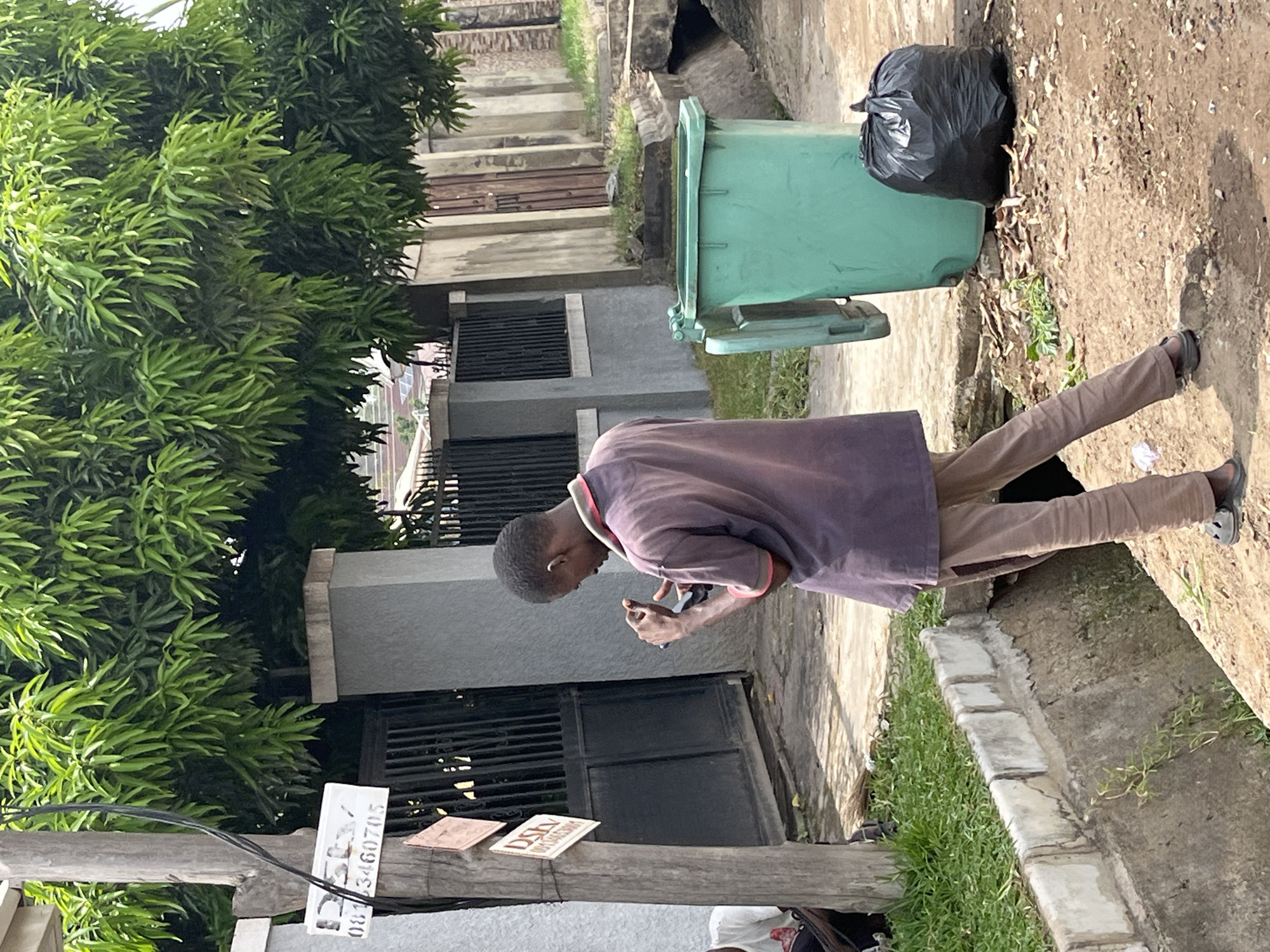 The scene is an outdoor setting in Abuja, Nigeria. The environment includes buildings with gray facades and barred windows, a lush green tree, and a concrete curb with grass growing nearby. A man stands on the sidewalk, focused on his mobile phone. He is wearing a purple shirt and khaki pants. Next to him is a green trash can and a black garbage bag. A utility pole with attached signs is also visible. One sign has a phone number, while another says Dstv.