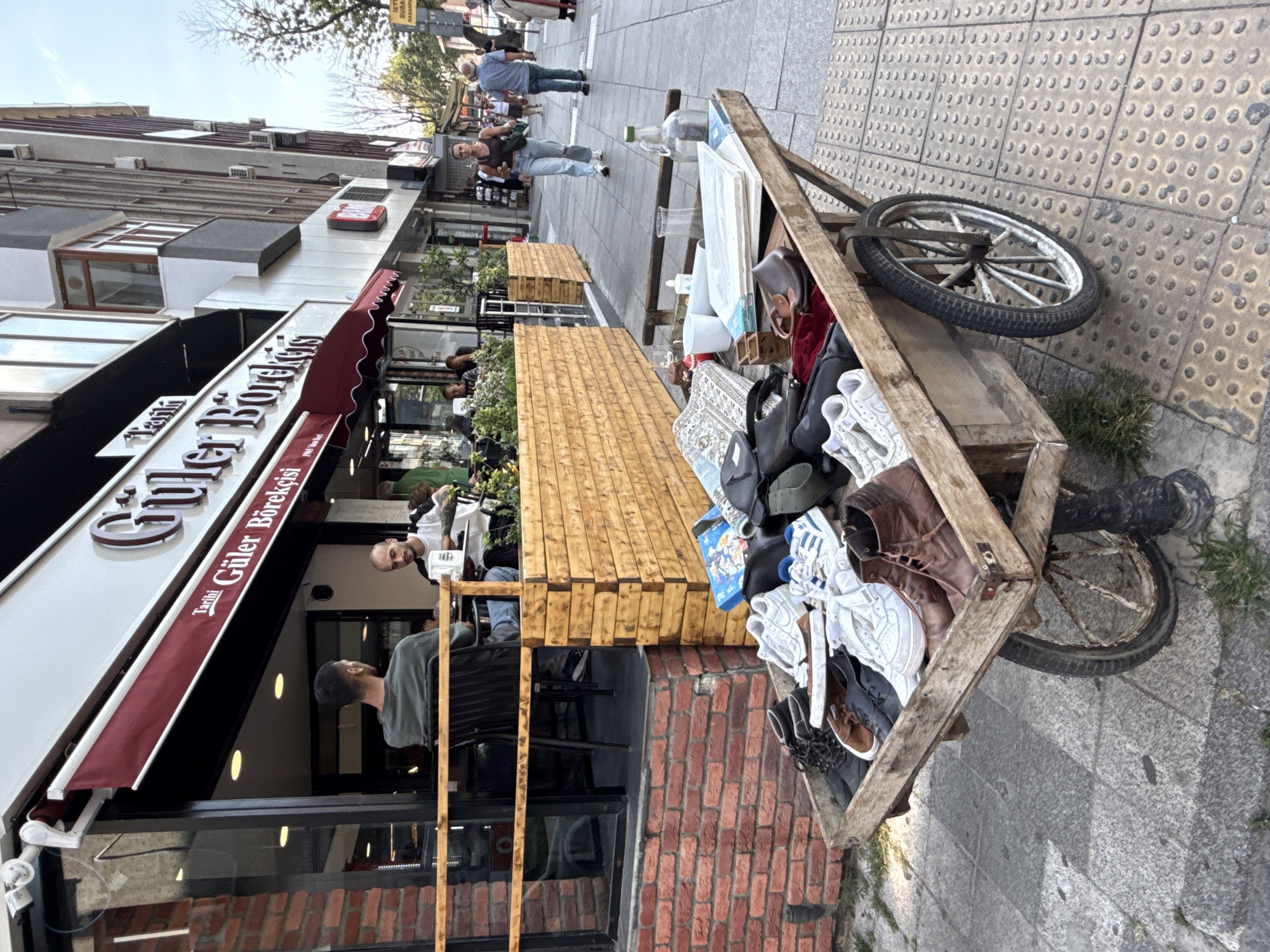 The image depicts a street scene in Kadıköy, Turkey, with a focus on a wooden cart laden with shoes and other items. The cart is positioned on a sidewalk made of textured grey tiles, with a brick wall and the exterior of a building to its left. The building appears to be a shop with large windows and a red awning. Inside the shop, two men are seated at a table.
The cart itself is a rustic wooden structure with two large wheels. It is filled with a variety of footwear, including sneakers, boots, and casual shoes. There are also some bags and other assorted items on the cart. The shoes are in various states of wear, suggesting they may be for sale or being transported.
In the background, pedestrians are walking along the sidewalk. The architecture of the buildings suggests an urban environment. The lighting indicates it is daytime. The overall impression is one of a busy street market or a vendor operating on the sidewalk.