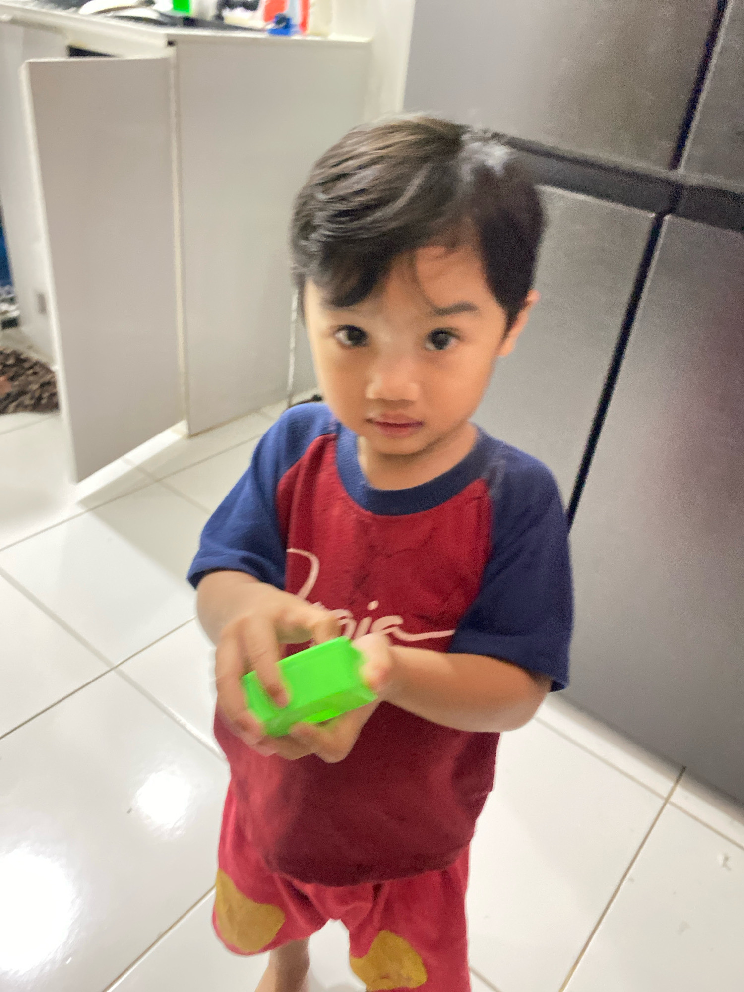 A young child, appearing to be between two and four years old, stands indoors in what appears to be a kitchen or utility area. The child has dark hair and is looking directly at the camera with a neutral expression. They are wearing a red t-shirt with blue sleeves and red shorts with yellow patches. The child is holding a small, rectangular green object in their hands.
The environment is characterized by white tiled flooring. To the left of the child, there is a white cabinet or refrigerator with a vertical seam. Further in the background, there are more white cabinets. To the right of the child, a large, dark grey appliance, likely a refrigerator, is visible. The lighting suggests an indoor setting, possibly with artificial light.
The image is taken from a slightly low angle, looking up at the child. The focus is primarily on the child, with the background being somewhat blurred. The location of Klurak, Indonesia, is not evident from the visual information present in the image itself; this detail would require external context.