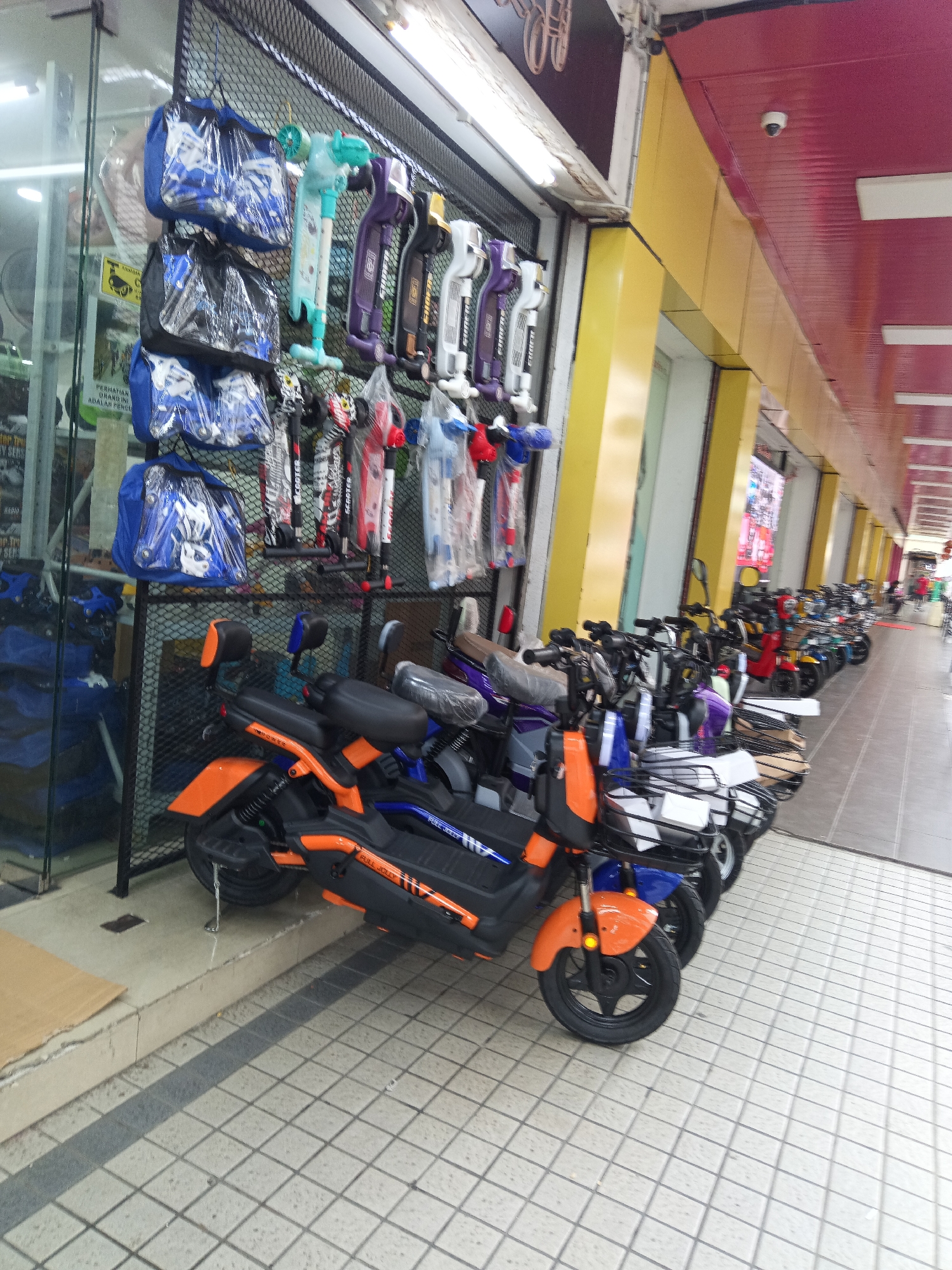 The image depicts a row of parked electric scooters and several scooters hanging on a wall outside a storefront. The storefront is a commercial establishment in Kuala Lumpur, Malaysia, characterized by its yellow and red facade. The scooters are predominantly orange and black, with some in blue and purple. Hanging on the wall are various bags, presumably for scooters, in blue. The ground is tiled with a grey and white checkerboard pattern. The scene appears to be a retail area, possibly a dealership or shop selling scooters. No people are clearly visible in the foreground, but a few individuals can be seen in the distance further down the street.