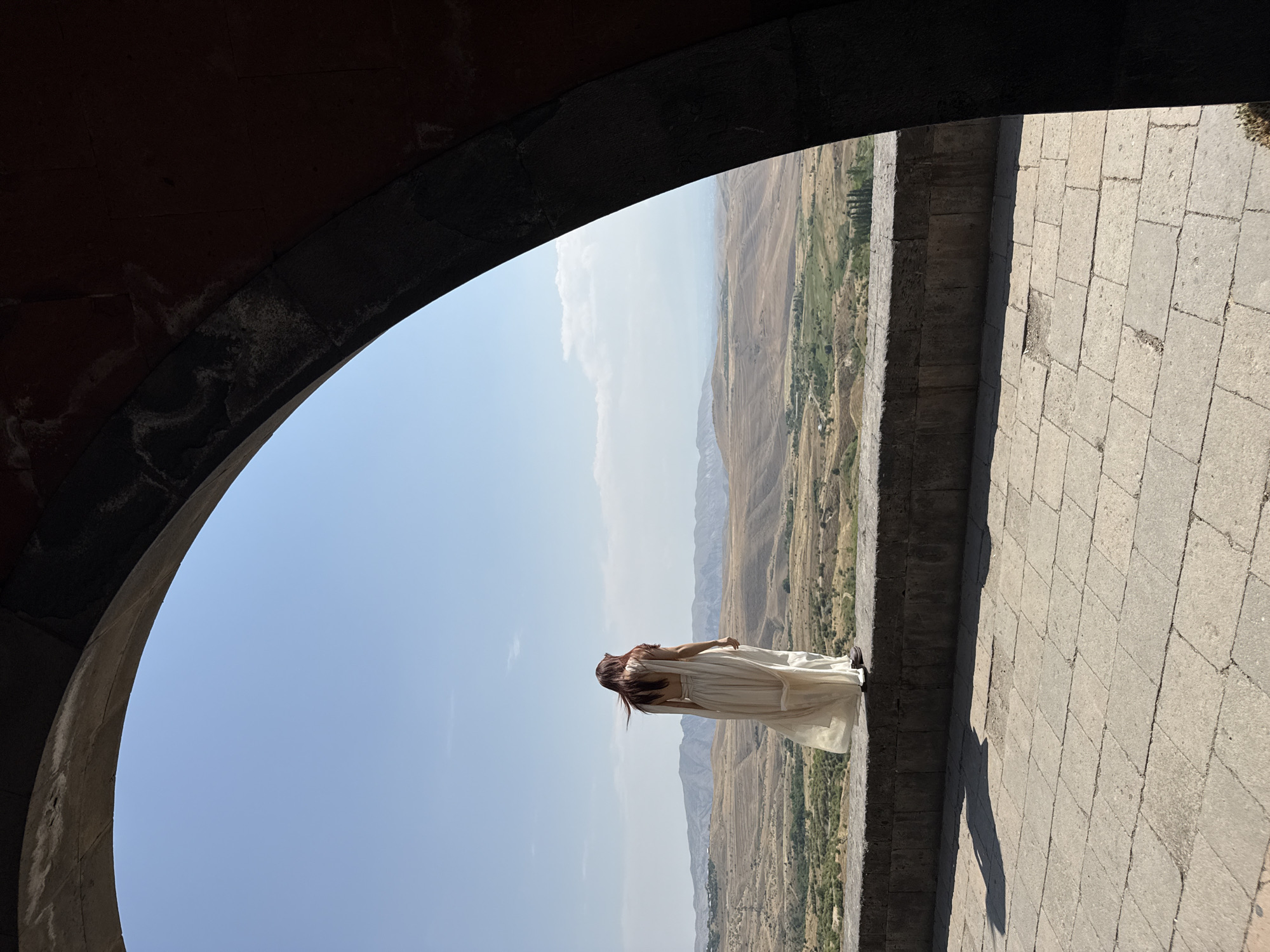 A person wearing a flowing white dress stands on a stone wall, looking out at a vast, hilly landscape under a bright blue sky. The view is framed by a dark, arched stone structure, suggesting they are within a building or a covered walkway. The landscape is arid, with brown and green hills stretching towards a distant mountain range. The ground in the foreground is paved with large, square stone tiles, casting a long shadow from the person and the wall. The scene appears to be in Voghjaberd, Armenia, a location known for its historic fortifications and scenic views.