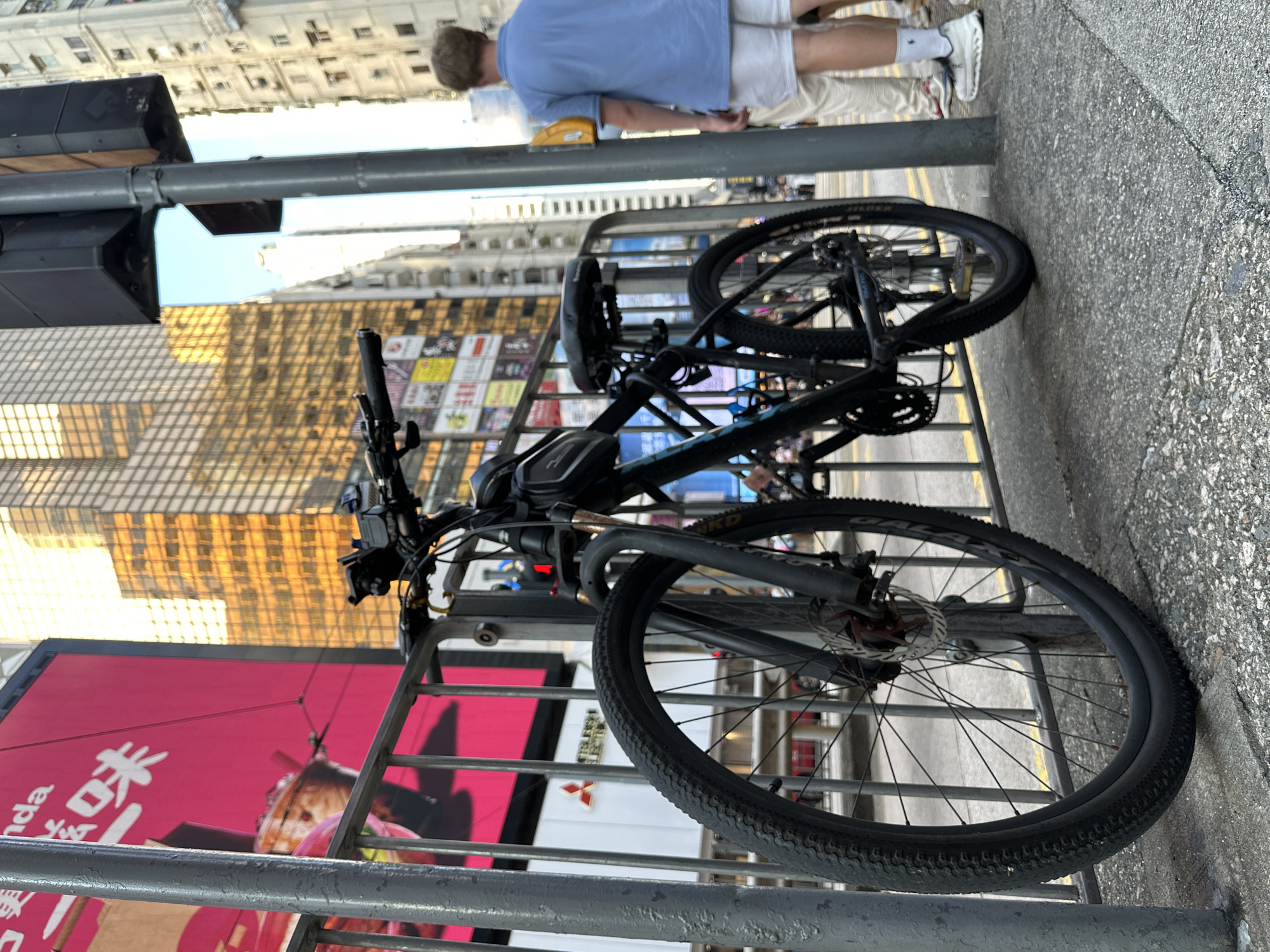 The image depicts a street scene in Hong Kong, China, focusing on a black bicycle parked next to a metal fence. In the background, a tall, gold-tinted building dominates the skyline, reflecting the sunlight. Adjacent to this modern skyscraper, older, high-rise buildings with multiple windows are visible. A bright pink billboard featuring Chinese characters and an image of a drink is positioned behind the fence. A traffic light pole stands to the right of the bicycle. A person wearing a blue polo shirt and white shorts is standing in the background, facing away from the camera, with their back to the street. The bicycle is a mountain bike with disc brakes and knobby tires. The ground is paved with concrete and asphalt.
