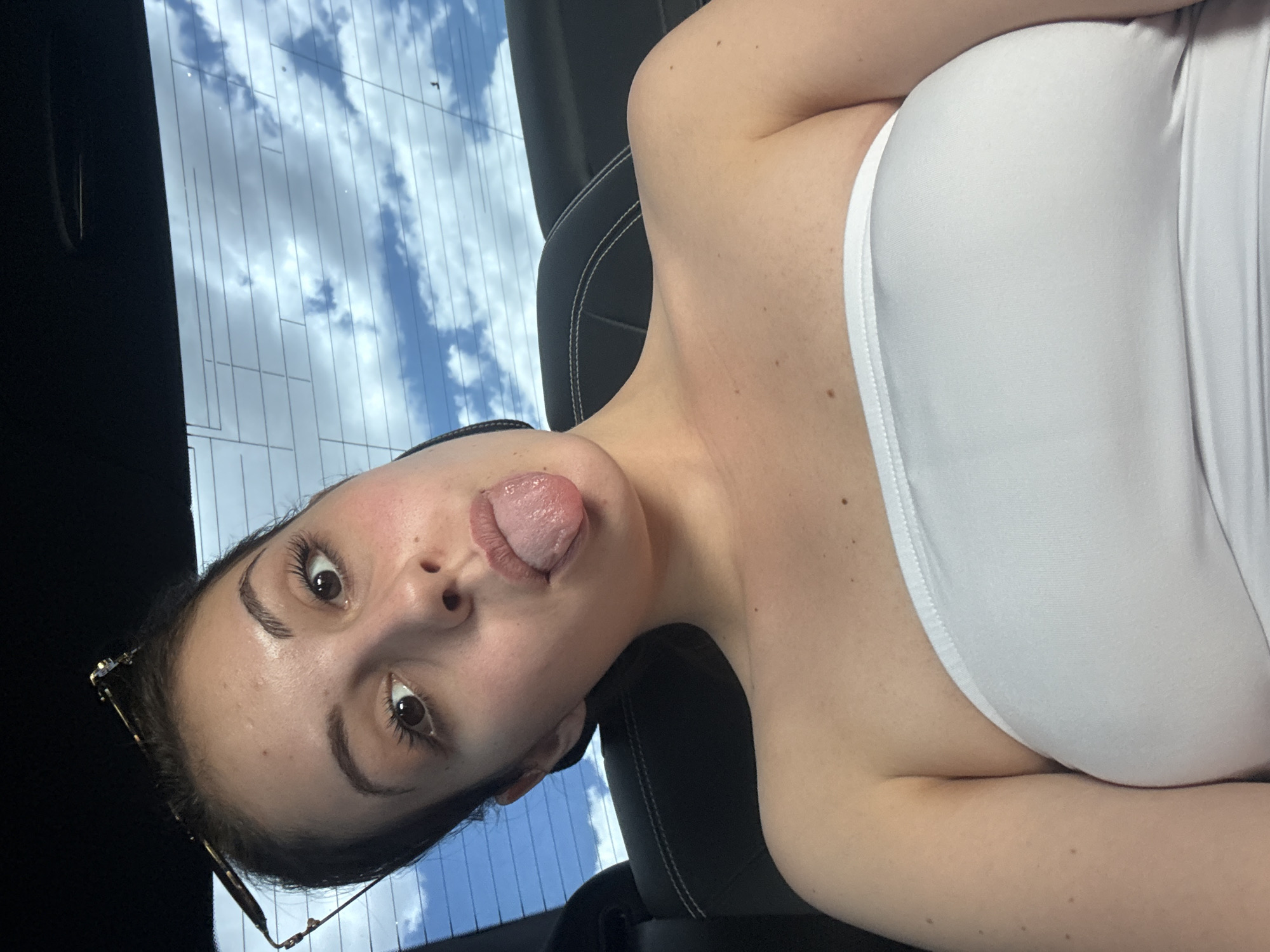 A young woman with dark hair, wearing a white strapless top, sticks her tongue out at the camera while sitting in a car. Her sunglasses are perched on her head. The background shows the sky with clouds through the rear window of the car.