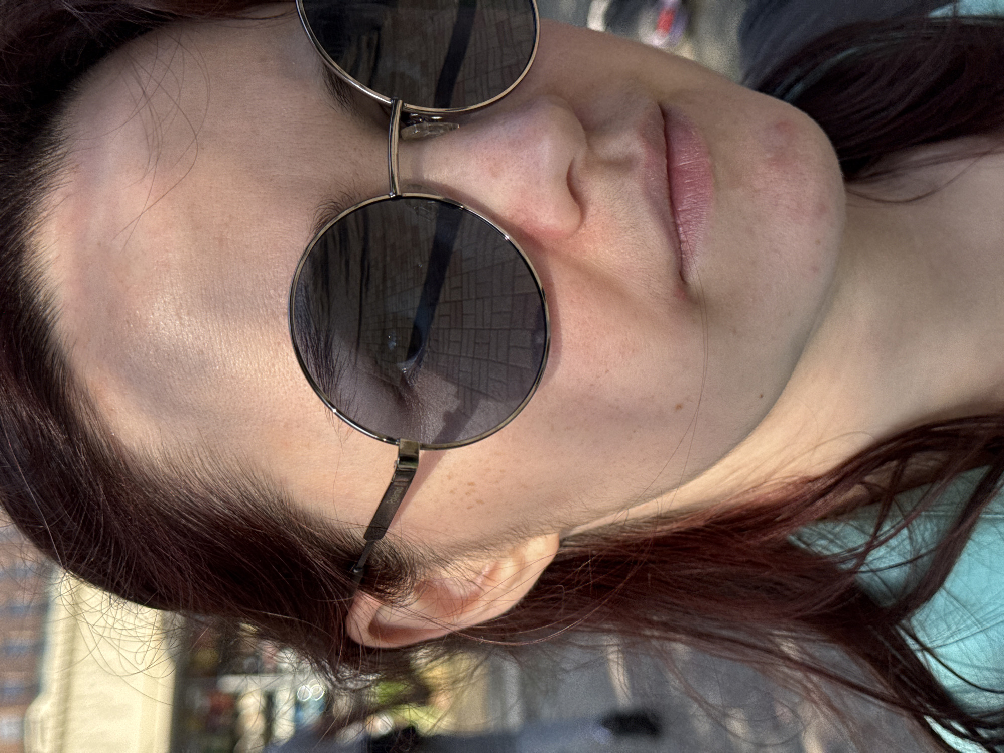 The image depicts a close-up of a person wearing round sunglasses. The glasses reflect a brick-paved area that suggests an urban outdoor setting. The subject has light skin, brown hair and is facing slightly off-center. The background, visible through the reflection and partially around the subject, suggests an outdoor urban environment. There is no specific indication in the image to identify the location as Sochi, Russia.