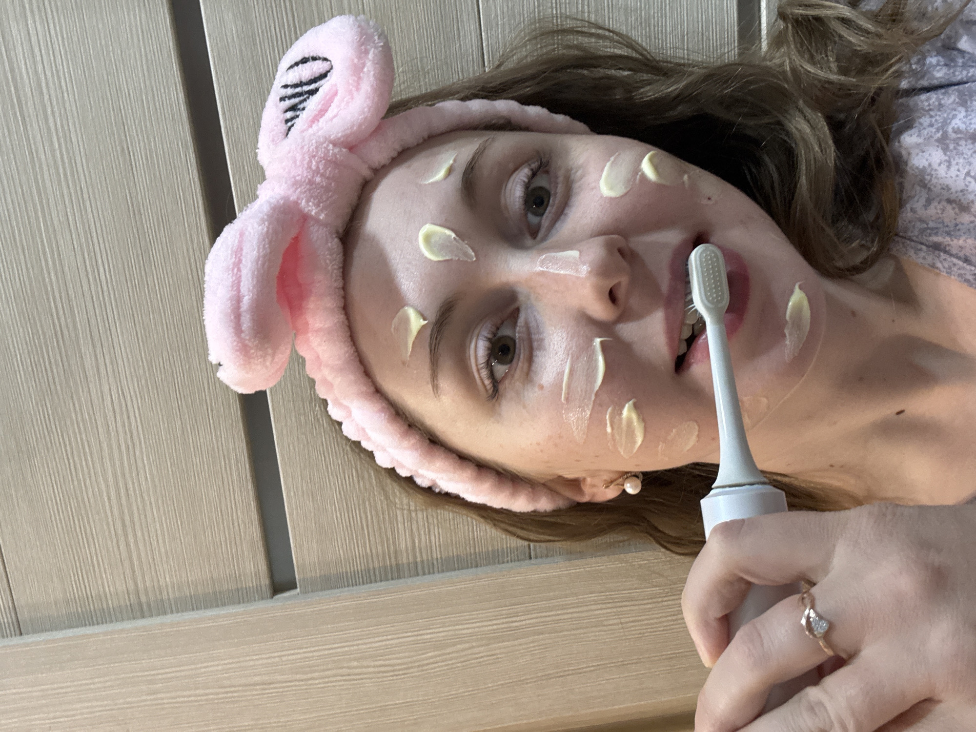 A woman with blonde hair is brushing her teeth while lying down. She has a pink headband with bunny ears and patches of a yellow substance on her face. She is holding a white electric toothbrush. The background consists of light brown wood paneling.