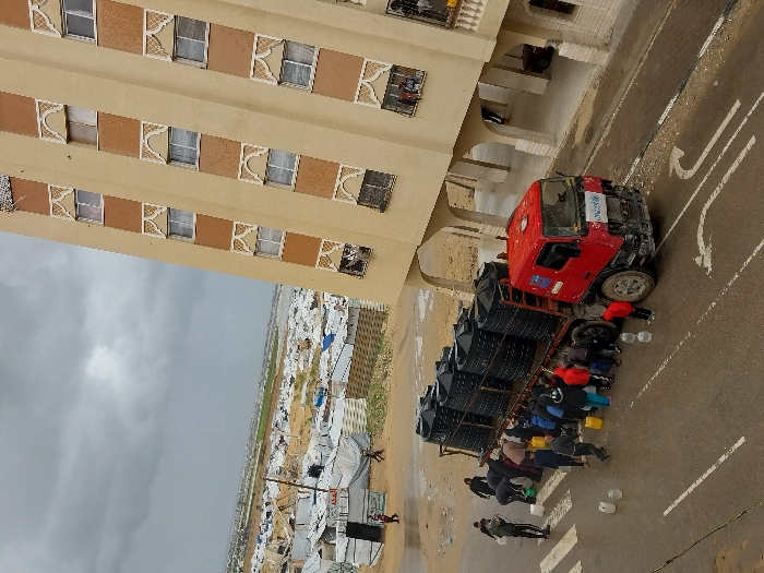 An overhead view captures a scene in Khan Yunis, Palestinian Territory, under an overcast sky. A red flatbed truck, marked with a blue UNICEF logo on its cab, is stationary on a wet asphalt road. The truck's bed carries approximately ten large, black, cylindrical water storage tanks. A group of about 15-20 individuals, including adults and children, are clustered around the truck's side, holding yellow and white jerrycans and buckets, appearing to collect water directly from the truck or its tanks. Several empty jerrycans are visible on the wet pavement.
To the right, a multi-story building with a light beige facade and vertical reddish-brown stripes stands. It features multiple windows with decorative, light-colored arched frames. The ground floor of this building appears open with arches, functioning as a covered walkway or entrance.
In the background, beyond the road and to the left of the building, a vast encampment stretches across sandy terrain. This encampment consists of numerous white tents and makeshift structures, indicating a large displaced population. Beyond the encampment, distant fields or natural landscapes are visible under the same cloudy sky. The road surface shows white painted markings for a crosswalk and a directional arrow.