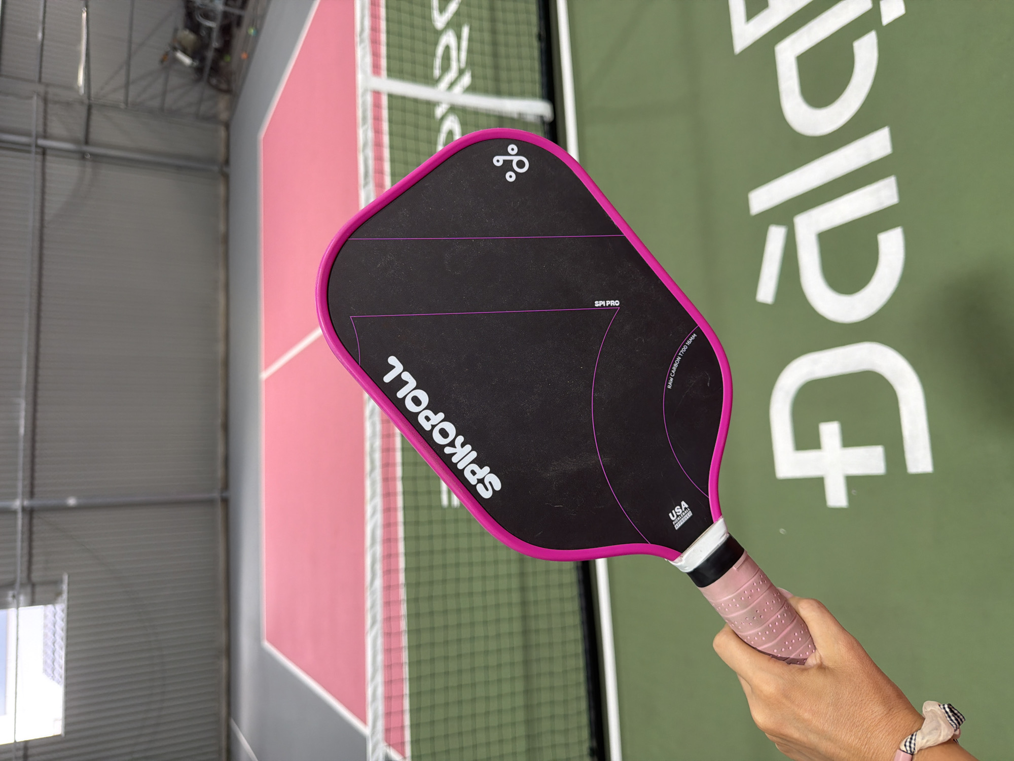 A person's hand is holding a pink and black pickleball paddle on a pickleball court. The paddle has "SPIKOPOLL" printed in white on the black surface. The court has a pink surface with white lines, and a green surface outside the court boundaries. A white net is visible in the background. The location is the city of Da Lat, Vietnam.