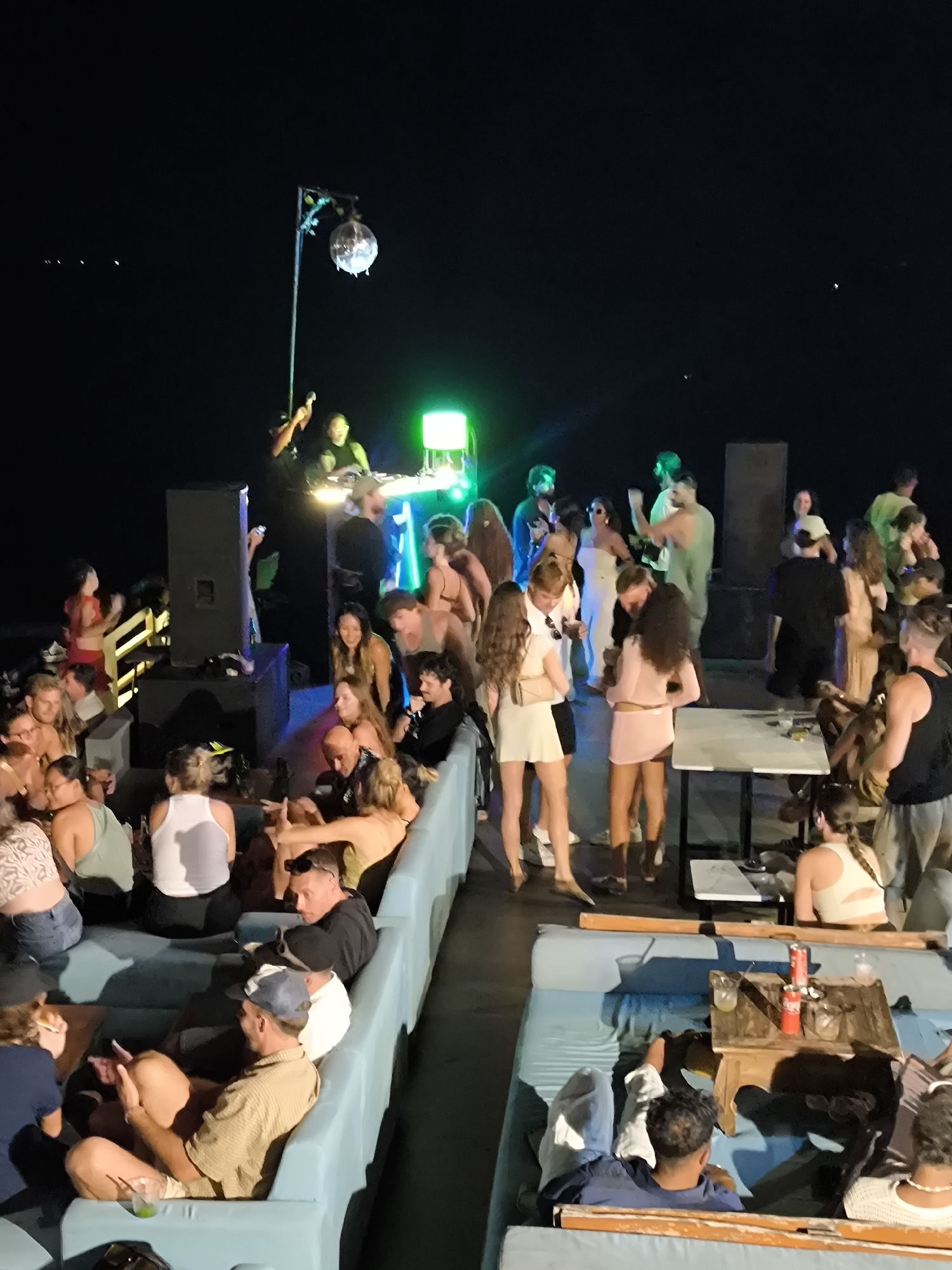 This is a high-angle nighttime photograph taken at a party in Uluwatu, Indonesia. A large group of people are socializing in a bar setting. A DJ is visible on an elevated platform with green lighting and a disco ball. The crowd is generally facing the DJ booth. Tables and lounge seating are occupied by patrons. The environment appears to be an open-air venue with dark sky visible.