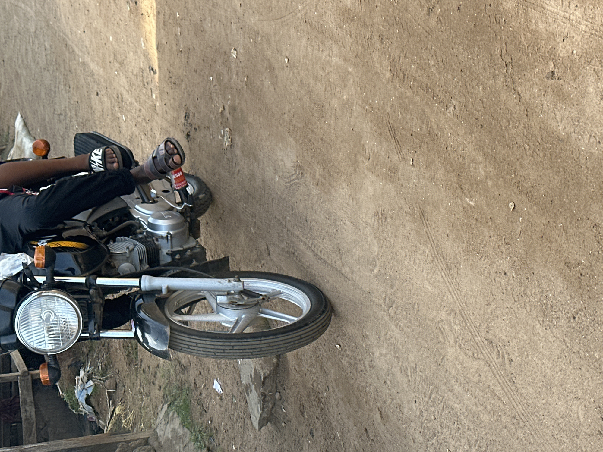 A low-angle, medium shot captures the front half of a black motorcycle with chrome accents, parked on a dusty, dirt road in Narayi, Nigeria. A person, dressed in black trousers and wearing black Nike slides on their left foot and brown sandals on their right, is seated on the motorcycle, their feet positioned on the footpegs. The motorcycle's headlight, orange turn signals, and front wheel are visible. The road surface is uneven and sandy, with tire tracks and scattered debris. In the background, a wooden structure and some dry vegetation are partially visible. The lighting suggests it is daytime, with soft shadows cast on the ground.