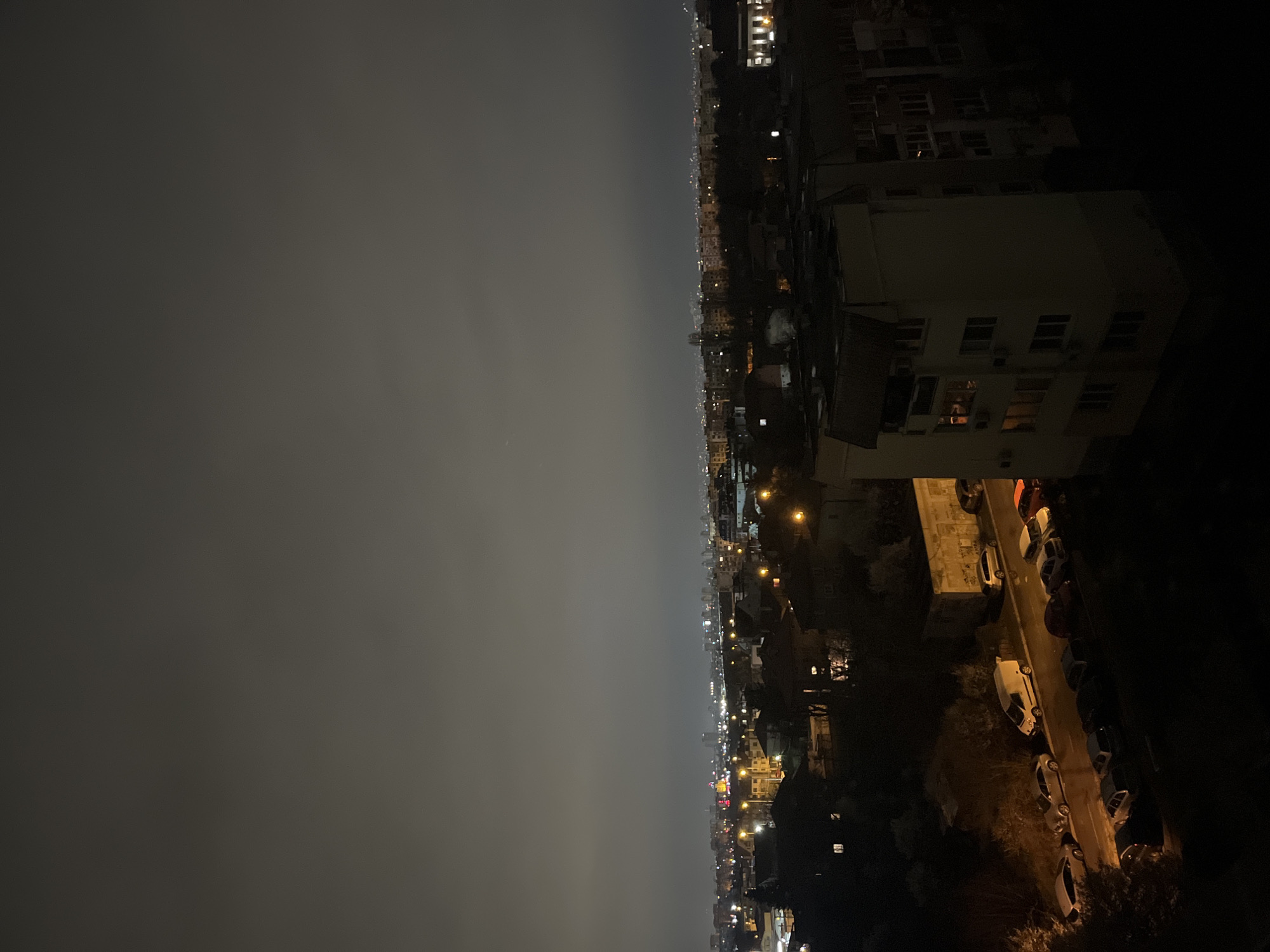 The image shows a nighttime view from what appears to be an elevated position overlooking a city, possibly Sofia, Bulgaria. The right side of the image captures a mix of buildings, from apartment blocks to smaller structures. A street is visible with parked cars. The left side of the image is dominated by a dark, cloudy sky. City lights illuminate parts of the landscape. There are no visible people or interactions.