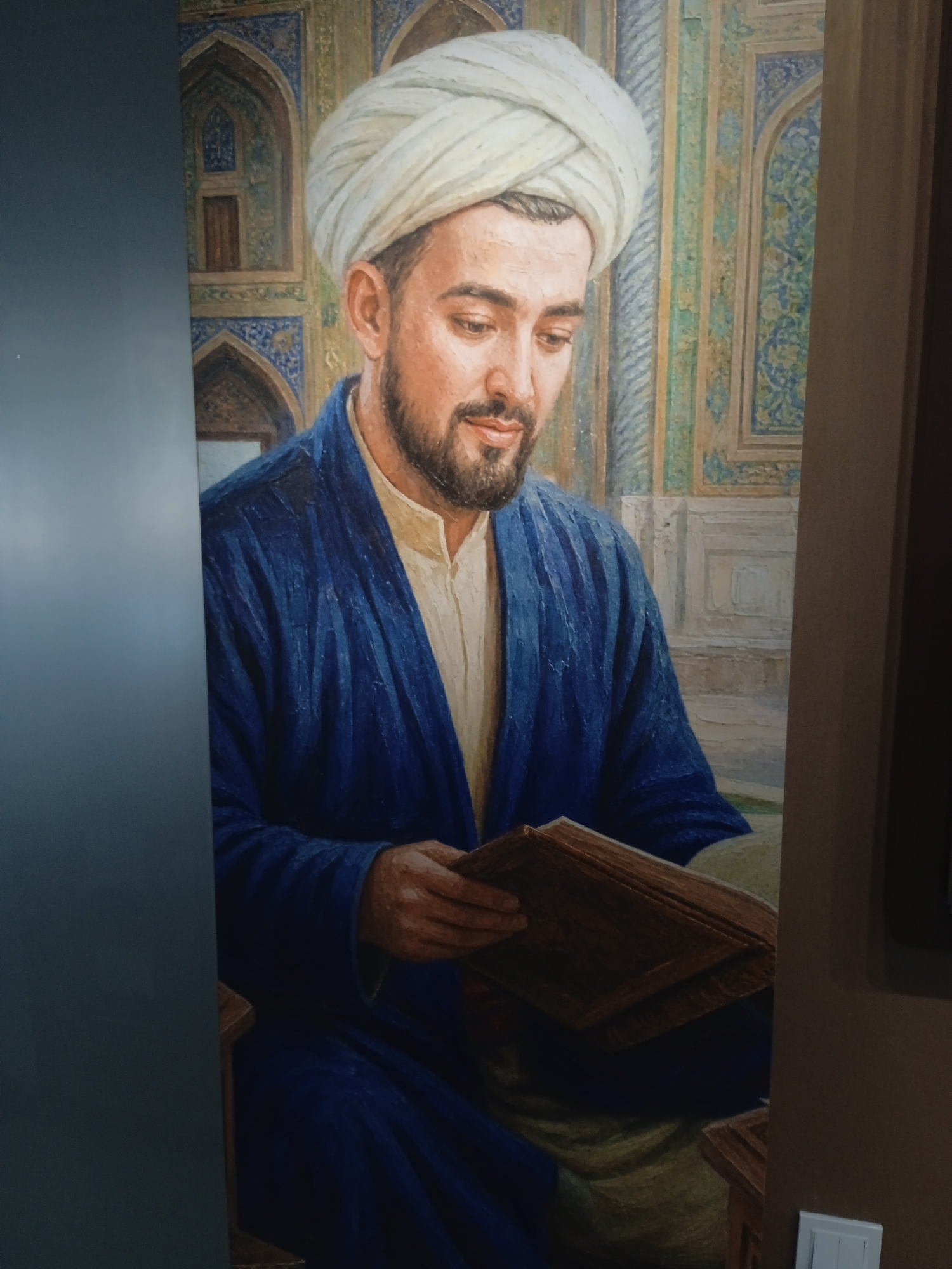 The image shows a portrait of a man wearing a white turban and a blue robe over a lighter-colored garment. He has a beard and is looking down at an open book in his hands. The background appears to be an interior with ornate, tiled walls featuring geometric patterns in shades of blue, green, and gold. The man is seated, and parts of what seem to be a wooden chair are visible. The lighting suggests an indoor setting. The photograph is taken from a slightly upward angle, with a dark, solid surface on the left side partially obscuring the portrait.