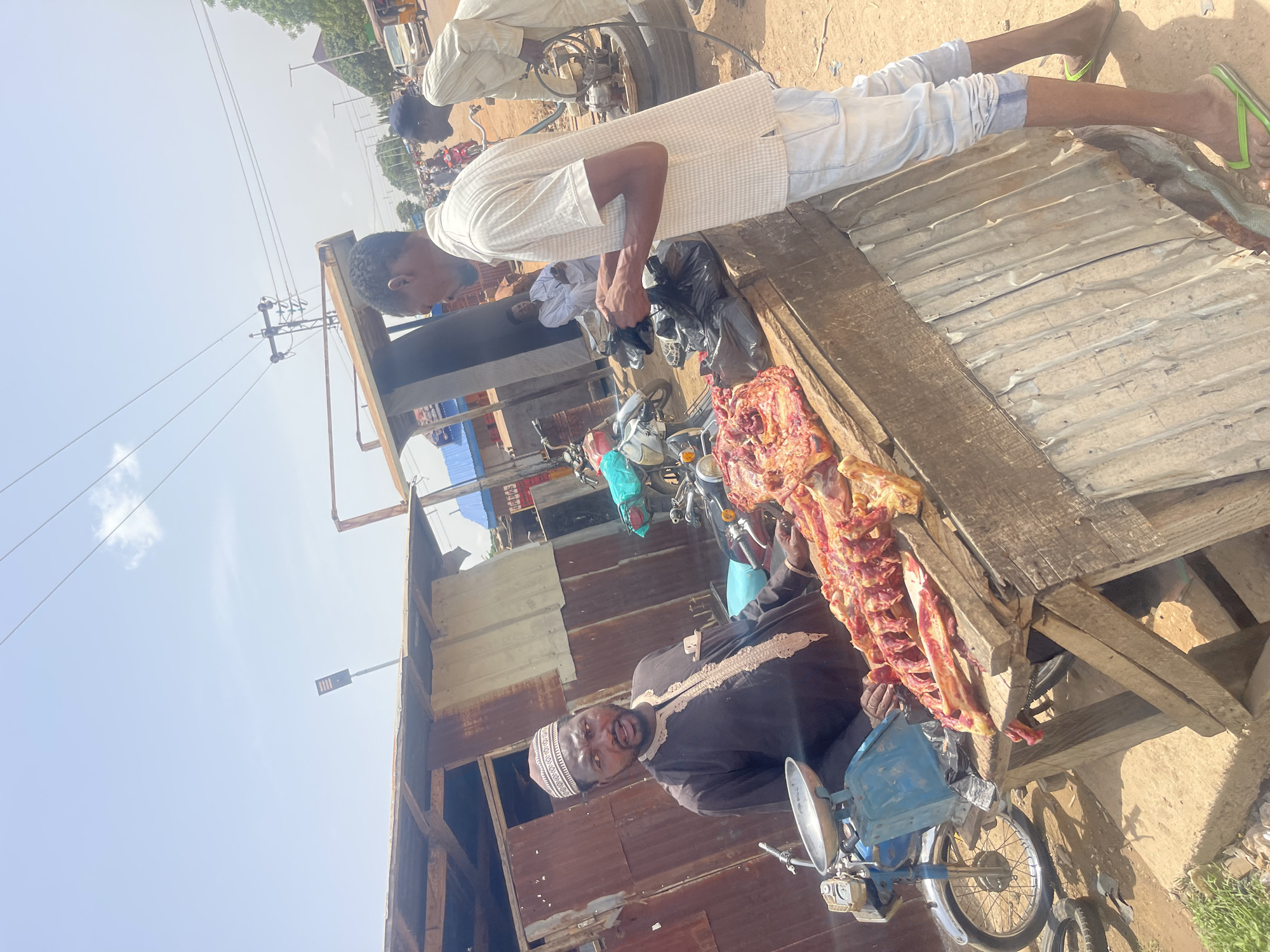 The image shows a street scene in Chikaji, Nigeria, where two men are engaged in the sale of meat. One man, wearing a dark robe and a white patterned hat, is seated on a blue motorcycle, likely the meat vendor. The other man, dressed in a white shirt and light pants, is standing by a wooden table, placing meat into a black plastic bag. A large piece of raw meat, complete with ribs and flesh, is displayed on the table. The surrounding environment includes corrugated metal structures, utility poles, other motorcycles, and a dusty ground, indicating a typical open-air market or roadside vending setup.