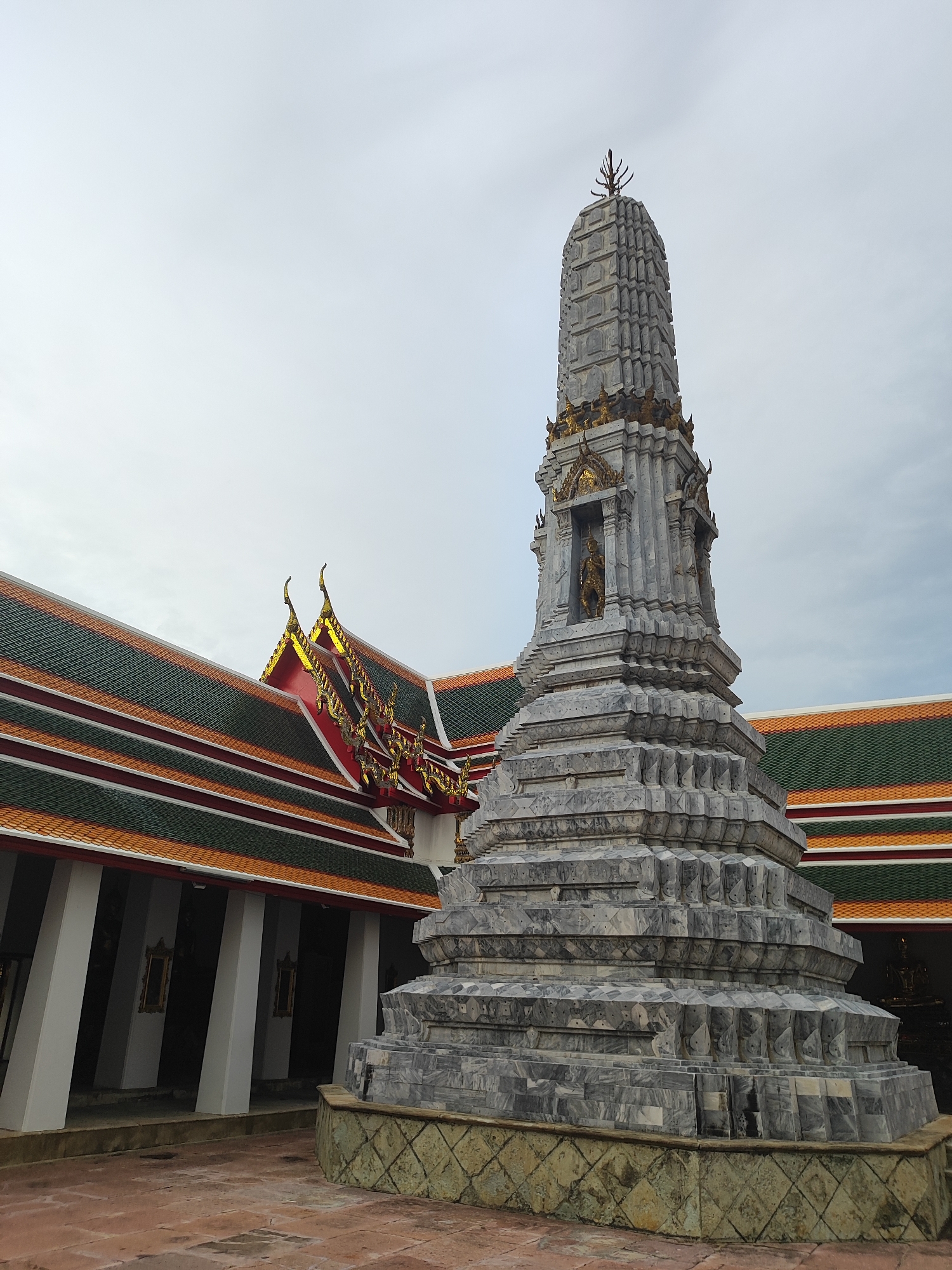 A grey, ornate stupa with gold accents stands in the foreground of the image, positioned to the right of center. The stupa is multi-tiered, with intricate carvings and statues visible at various levels. Behind the stupa, to the left, is a building with a traditional Thai architectural style, characterized by a steeply pitched, multi-layered roof covered in green and orange tiles, with ornate gold trim along the edges. The building has a covered walkway supported by white pillars. The ground in the foreground is paved with reddish-brown tiles. The sky above is overcast and a pale blue-grey. The image appears to have been taken at Wat Pho in Bangkok, Thailand, a prominent Buddhist temple complex. There are no people or animals visible in the scene.