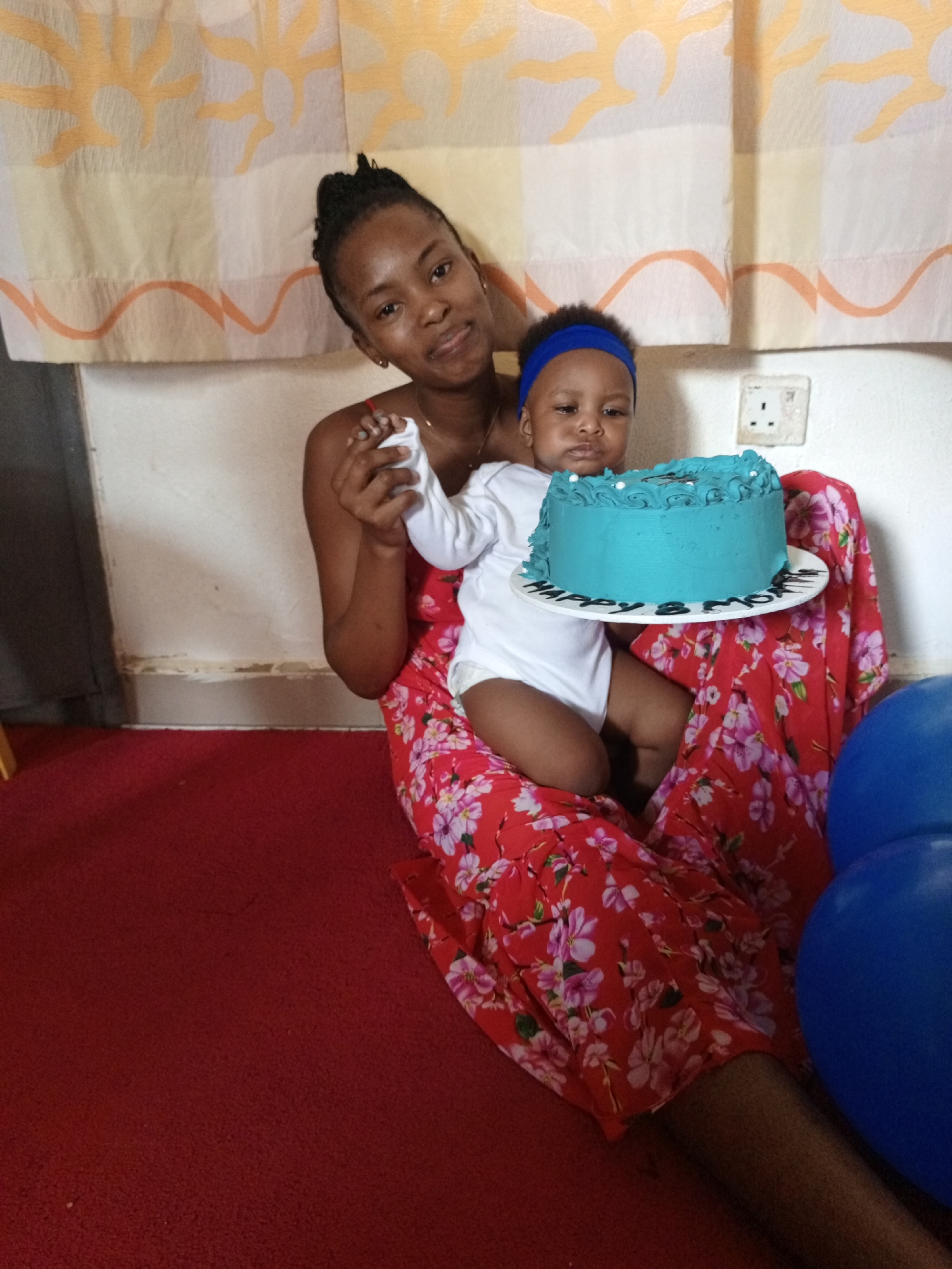 A woman and a baby are sitting on a red carpet, in an indoor scene in Keffi, Nigeria. The woman has her hair in a braided updo, and is wearing a red dress with a floral print. She is holding the baby, who is wearing a white onesie and a blue headband. Between them is a blue cake, decorated with white frosting and placed on a white plate that reads "Happy 8 months". To the right, in the corner, is a stack of blue balloons. The background includes a wall with orange and beige patterned curtains and a white electrical outlet. The scene appears to be a celebration of the baby's 8-month milestone.