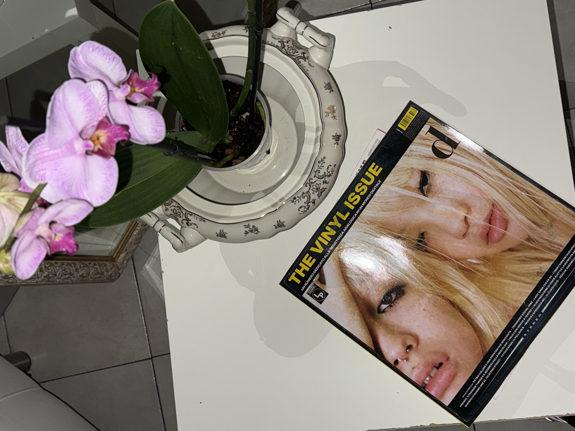 A high-angle indoor shot depicts a white, square table with several items on it. To the left is a purple orchid plant in a white ornamental planter set on a patterned base. To the right is an Italian magazine titled "The Vinyl Issue" with an image of two blonde women on the cover. The text is partially visible in Italian. The location is possibly in Sanremo, Italy, though this is not explicitly stated.