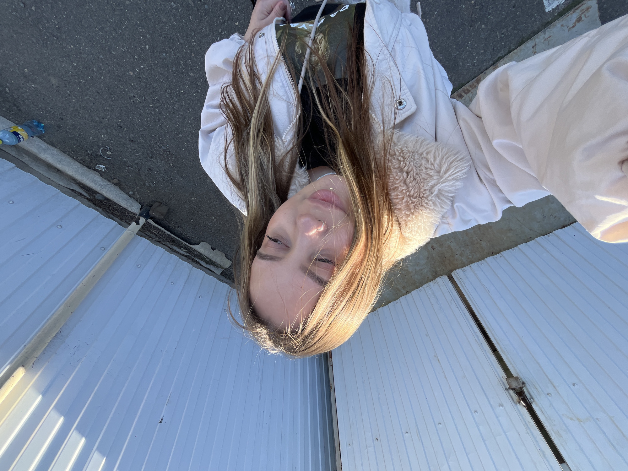 A young woman with long, light brown hair and a nose ring is looking into the camera. She is wearing a black shirt, a light-colored jacket with a fur-lined collar, and appears to be standing near a corrugated metal fence or wall. A portion of a concrete surface, potentially a sidewalk or road, is visible in the background. Debris, including a bottle, is near the upper-left corner of the image. The photograph was presumably taken in Novopokrovskaya, Russia.