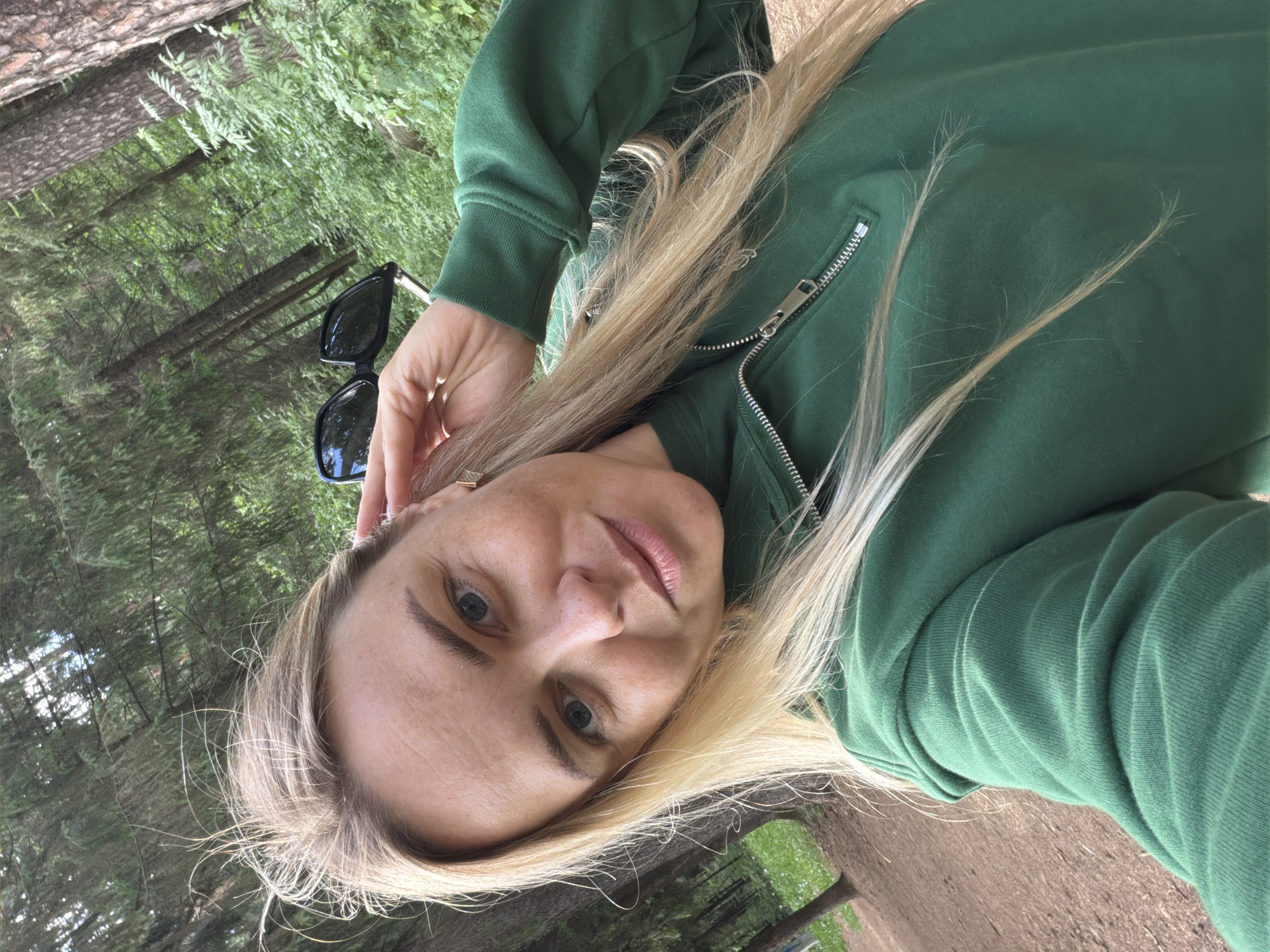 A woman is pictured from a low angle outdoors. She has fair skin, light blonde hair, and light-colored eyes. She is wearing a green zip-up jacket. Sunglasses are perched on top of her head. The background consists of a forest environment with trees and greenery. The exact location in the city of Votkinsk, Russia is not discernible from the image.