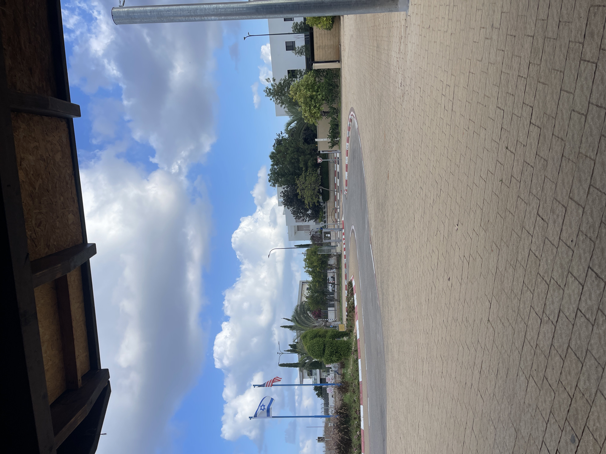 The image shows a paved roundabout with red and white curbs. Two flags, the Israeli flag and the American flag, are visible on flagpoles to the left of the roundabout. Behind the roundabout, there are buildings, trees, and greenery. The sky is blue with white clouds. The foreground is dominated by a stone-paved walkway. There are no people visible in the image. The scene appears to be a public area within the city of Even Yehuda, Israel, possibly near an official building or an embassy, given the presence of the two flags.