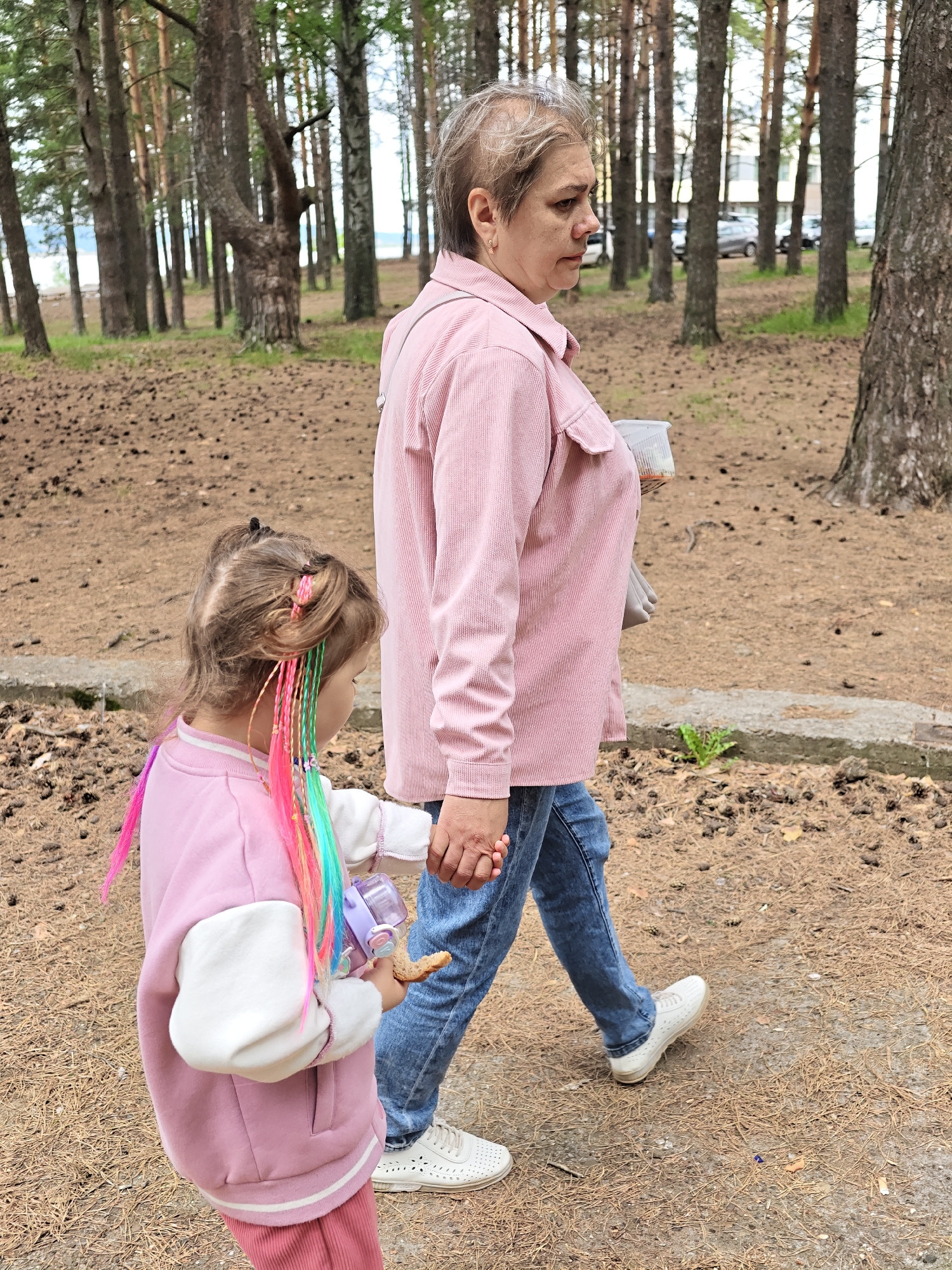 In a wooded area on the outskirts of Chaikovsky, Russia, an adult woman and a young child are walking on a path covered with pine needles and fallen cones. The woman, with short graying hair, wears a light pink corduroy jacket and blue jeans. She is holding a clear plastic cup with a white lid in her right hand. The child, facing away from the camera, wears a pink jacket with white sleeves and pink pants. The child's hair is styled in two pigtails with colorful extensions in pink, green, and blue. The child is holding a purple water bottle in their left hand and a piece of food in their right hand. The background consists of tall pine trees and a glimpse of a body of water and buildings in the distance.