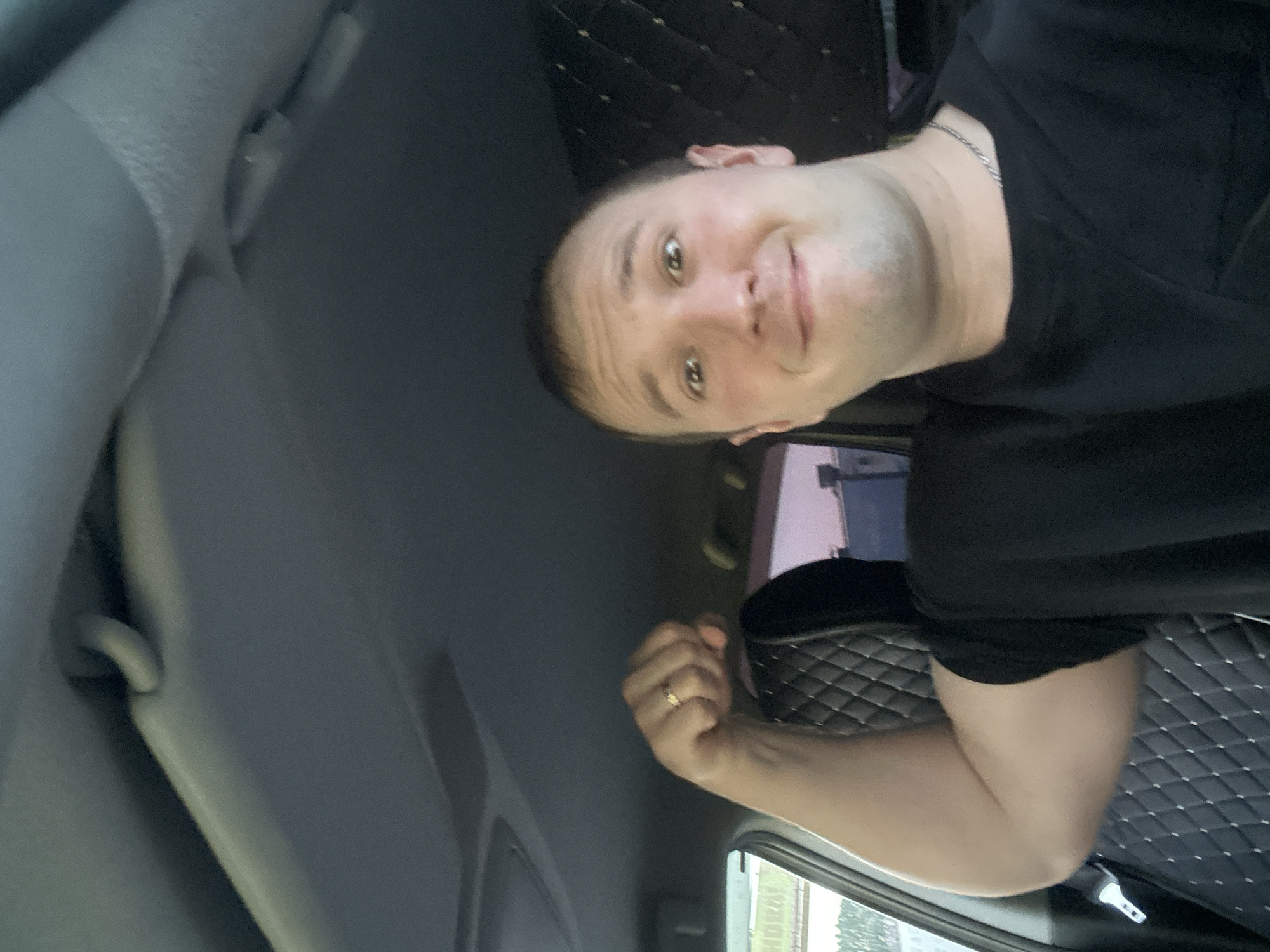 A selfie photograph, oriented at an angle, depicts a fair-skinned man in the interior of a vehicle. The man, wearing a black t-shirt and a thin silver chain, has short brown hair and is looking directly at the camera with a slight smile. He has a gold ring on his left ring finger and his left arm is bent. The vehicle's interior features grey upholstery on the roof and panels, as well as a black diamond-patterned cushion on the floor. Outside the vehicle's window, foliage and buildings are partially visible, indicating an urban or suburban environment. Although the location is stated to be Chaikovsky, Russia, this cannot be forensically confirmed from the visual data alone.