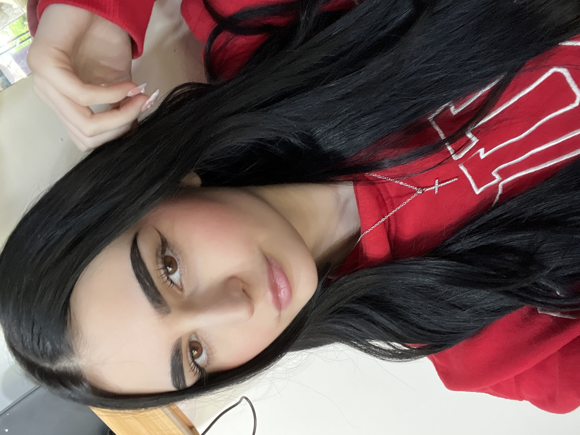 A young woman with long, dark hair is lying down, wearing a red hooded sweatshirt with white trim. She is fair-skinned with brown eyes and defined eyebrows. She is wearing a silver necklace with a cross pendant. Her fingernails are long and manicured. The background is blurred and indistinct. The location, Burgas, Bulgaria, is not discernible from the image.