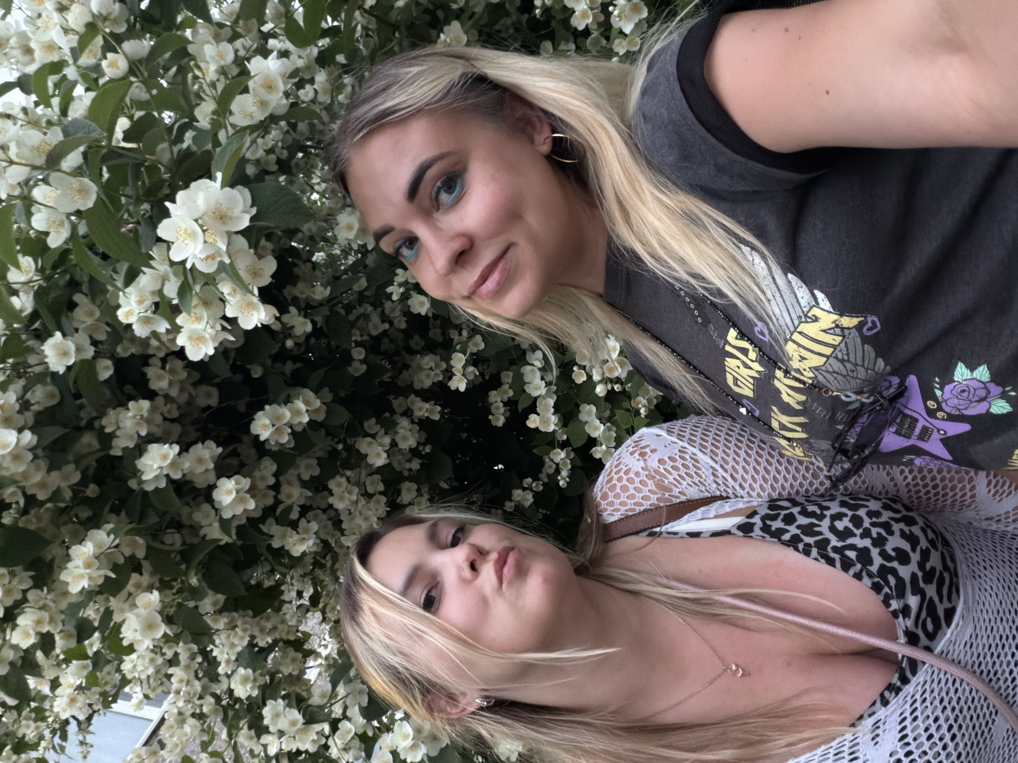 Two young women are posing for a selfie in front of a dense bush covered in white flowers, likely jasmine, possibly in a park or garden in Chaikovsky, Russia. The woman on the left, with blonde hair and a leopard print top under a white mesh cover-up, puckers her lips towards the camera. The woman on the right, also with blonde hair and bright blue eyeshadow, smiles slightly and tilts her head. She is wearing a dark t-shirt with a graphic print. The background is dominated by the white blossoms and green foliage of the bush.