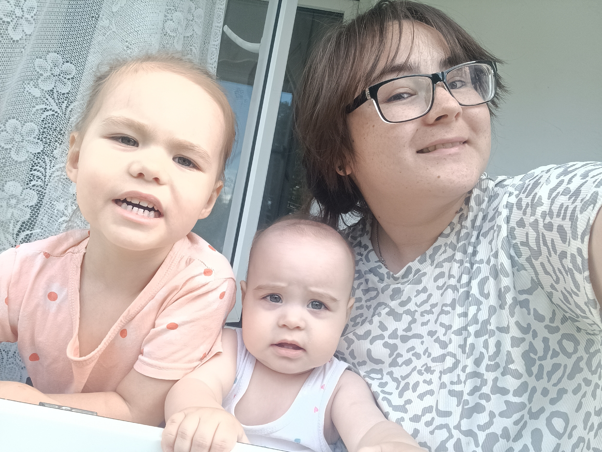 The image shows a selfie featuring three people, likely a mother and her two children, outdoors in what appears to be a residential setting. The woman on the right, wearing glasses and a patterned top, is smiling and looking at the camera. Next to her, a baby in a white tank top with a faint heart pattern is also looking forward with a neutral expression. On the left, a young girl with a peach-colored polka-dot shirt leans forward, her mouth open in a playful expression. A white lace curtain is visible behind the children, and a window frame suggests they are near a doorway or window. The overall lighting and slight overexposure suggest it is daytime, likely sunny. The environment appears to be casual and domestic, possibly a porch or balcony in Noviy, Russia. No visible text is present in the image.
