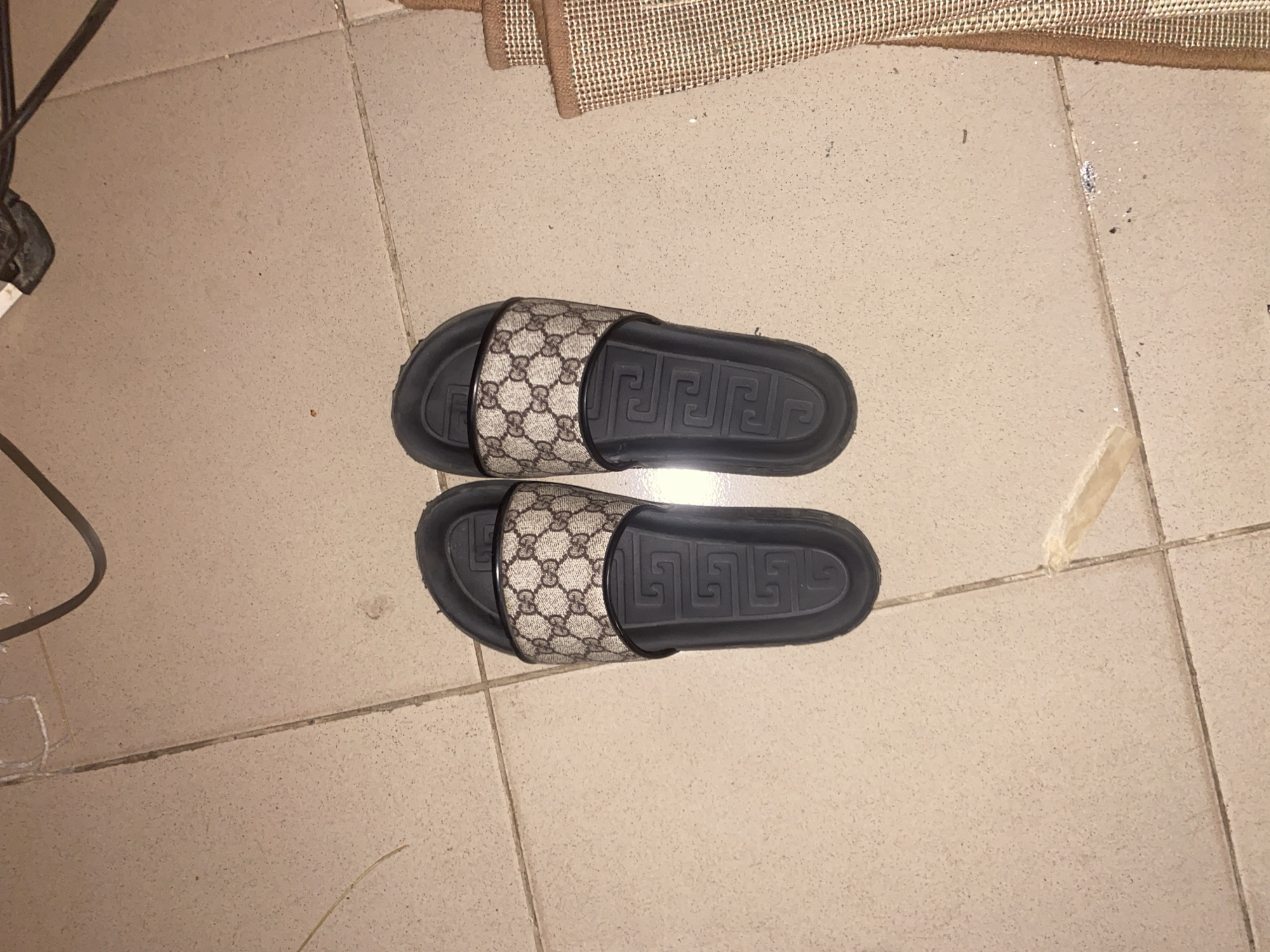 A pair of sandals are positioned centrally on tiled flooring. A rug is partially visible in the upper right. A cable and part of a mechanical device are visible in the upper left. The location is unknown.