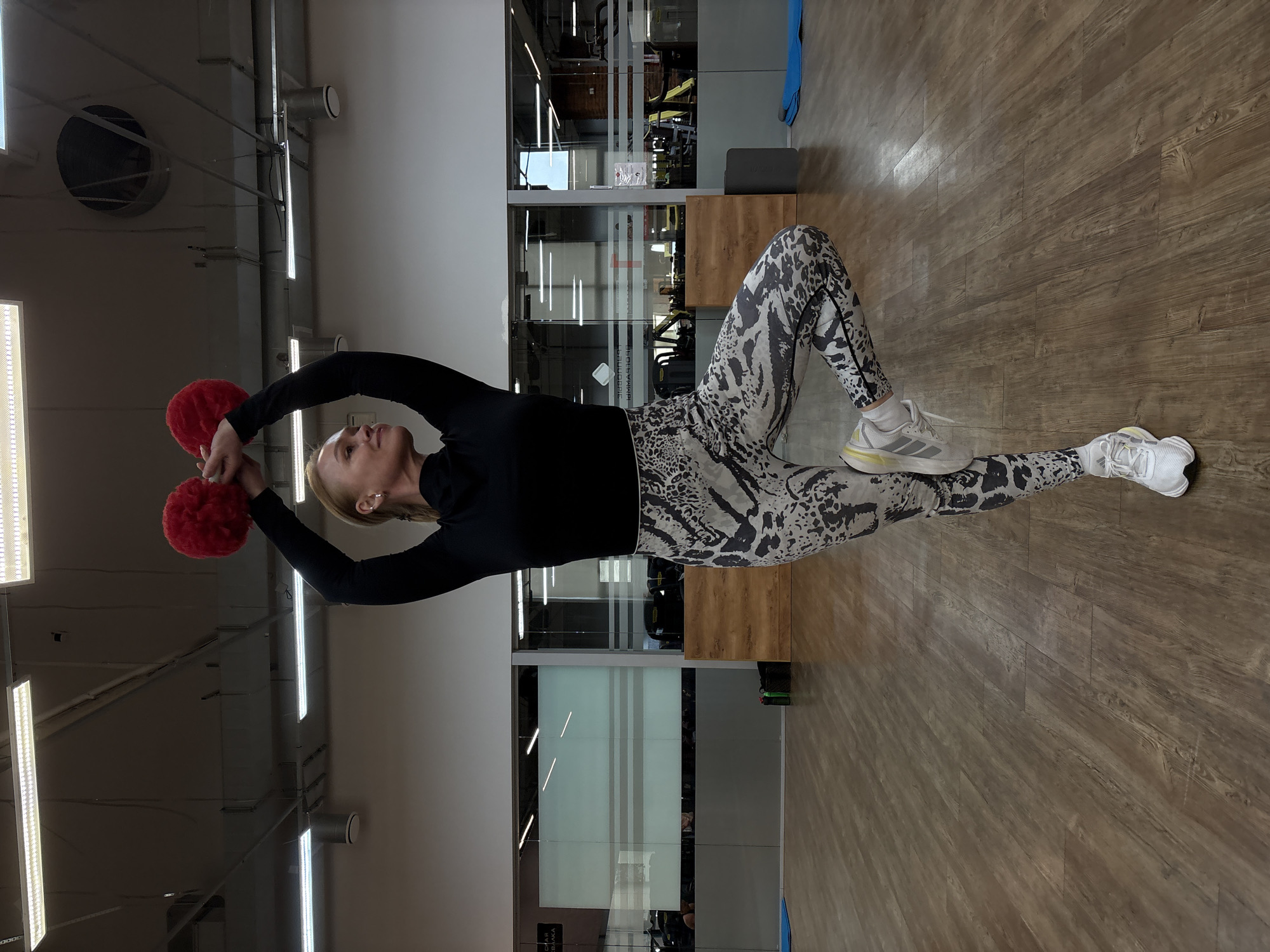 A woman is lying on a wooden floor in a gym in Chaikovsky, Russia. She is wearing a black long-sleeved shirt and patterned leggings, as well as white athletic shoes. She is holding two red, pom-pom-like objects above her with both arms. The gym contains exercise equipment and glass-walled rooms. Overhead, there are industrial light fixtures and ductwork.