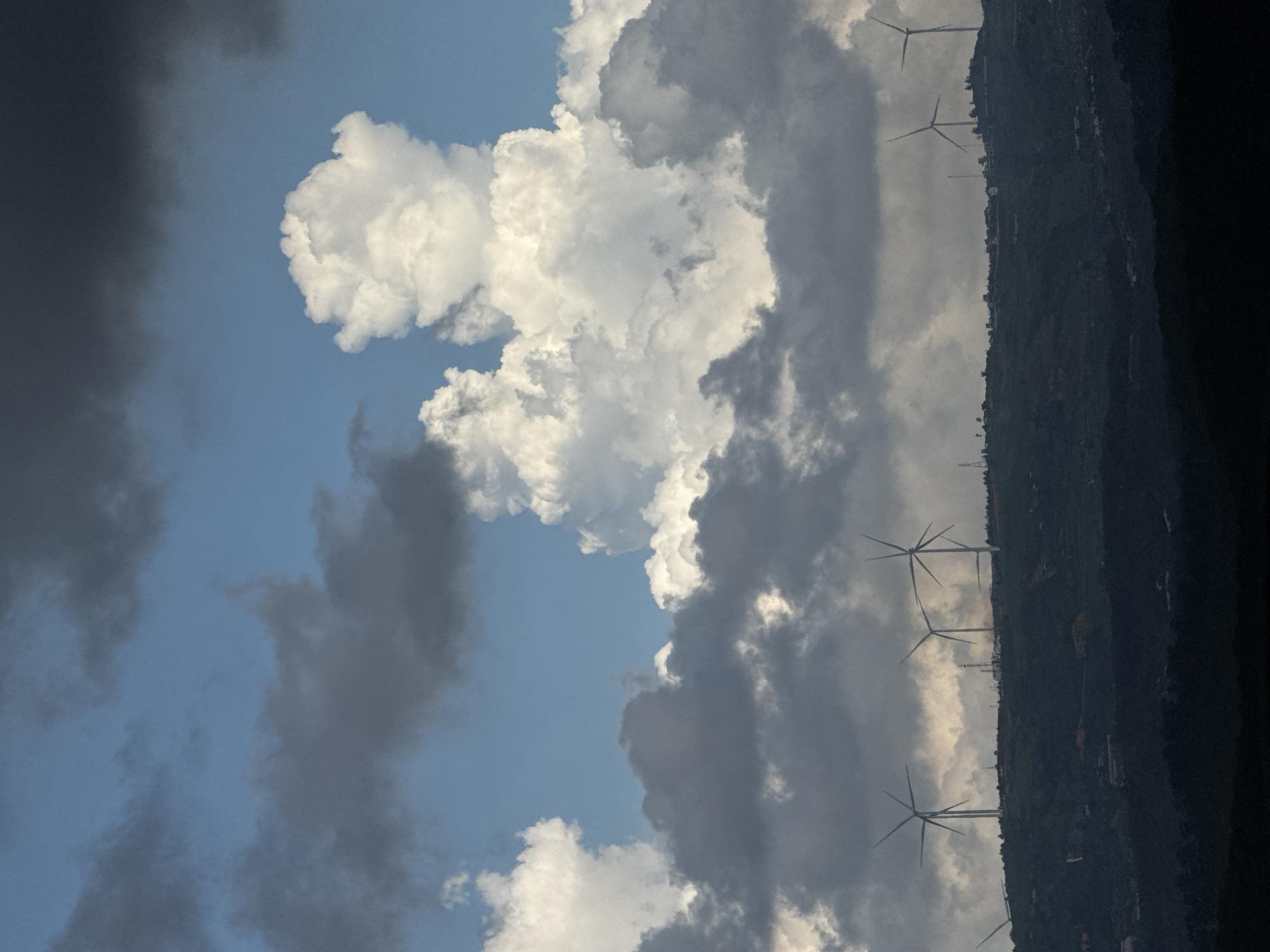 The image depicts a hilly landscape under a dramatic sky. Several wind turbines are visible along the ridge of a dark green, vegetated hill, silhouetted against a backdrop of clouds. The sky is a mix of deep blue, dark gray storm clouds, and bright white cumulus clouds, with the sun illuminating some of the cloud formations. The foreground is a dark, indistinct mass of land, possibly a lower hill or valley. The scene suggests a location with renewable energy infrastructure in a natural environment. The specific location in the city of Đa Lộc, Vietnam is not discernible from the image itself. There are no people or discernible actions taking place.