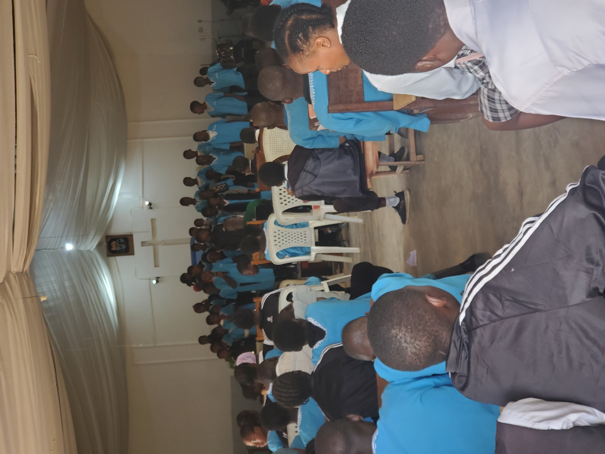 The image shows a group of people, primarily young individuals, seated in a room. They are mostly wearing light blue shirts with white collars, suggesting a uniform or group attire. Some are also wearing white shirts, and one person in the foreground is wearing a black jacket with white stripes on the sleeves. The individuals are seated on chairs and some are gathered around tables, with several people facing away from the camera.
The scene appears to be indoors, with a curtain or draped fabric on the left side of the image, possibly part of a stage or decorative element. There is a cross on the wall in the background, and a framed image with a number "19" inside. The lighting suggests it might be daytime, but the exact time is not discernible. The overall mood seems to be one of gathering or a formal event, possibly a religious service or a school assembly, given the presence of uniforms and the cross. The individuals are looking down or are in profile, making it difficult to discern specific emotions, though some appear engaged. The location cues, such as the cross and the architecture, might suggest a church or a school setting in Kuru Karama, Nigeria.