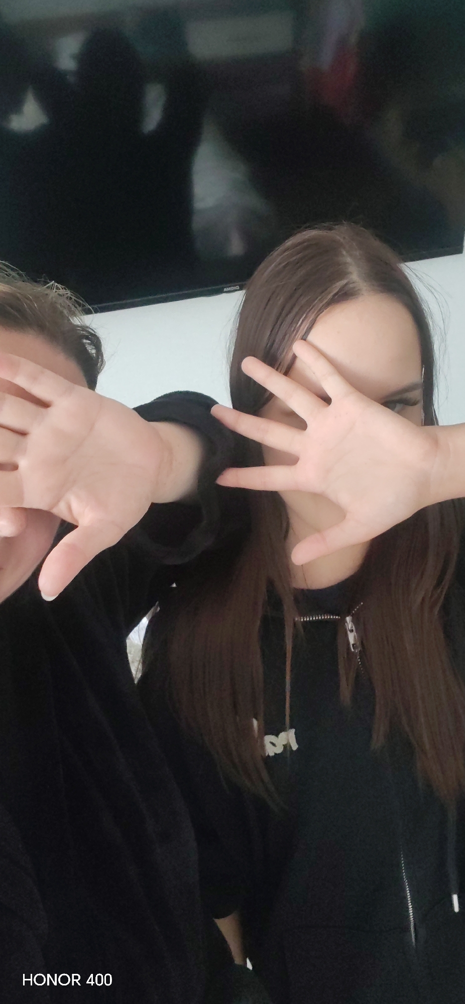 Two individuals are positioned in the foreground, partially obscuring their faces with their hands. The person on the right has long, straight brown hair and is wearing a black zip-up hooded sweatshirt with a logo on the chest. The individual on the left is also wearing black clothing. In the background, there is a television screen reflecting a shadowy figure. The lower left corner of the image has the text "HONOR 400". The location, Noviy, Russia is not determinable from the image.
