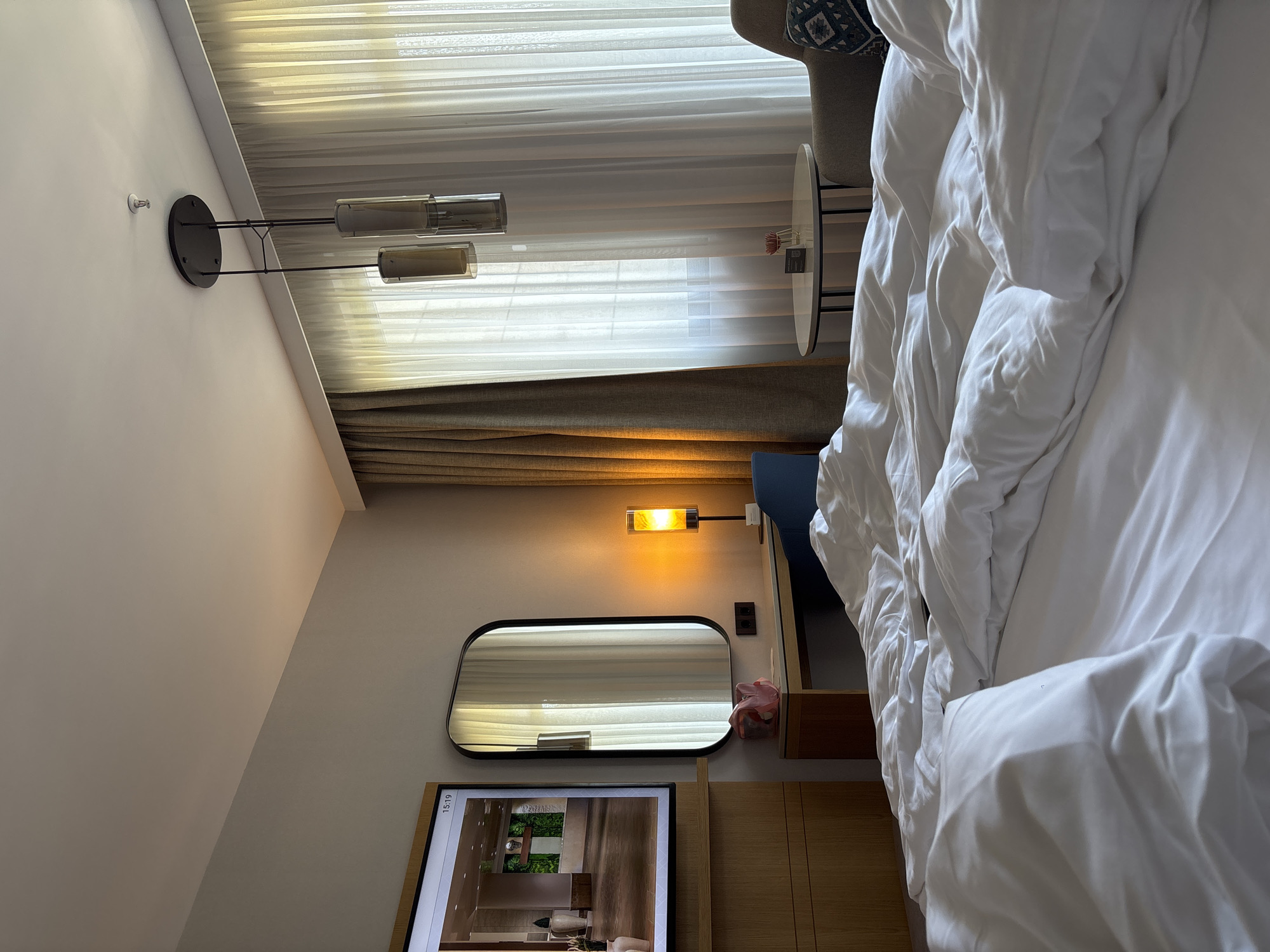 The image depicts a hotel room in Razliv, Bulgaria. A rumpled bed with white linens occupies the foreground, suggesting recent occupancy. To the left, a large television screen displays an image of a modern interior. Adjacent to the television is a wooden dresser. Further left, a wall-mounted mirror reflects the room and a window with sheer, white curtains. A desk with a warm-toned lamp, a blue chair, and a pink object sits beneath the mirror. In the background, a small circular table with a vase holding a single red flower and a small card is visible next to an armchair. A modern pendant light fixture hangs from the ceiling.