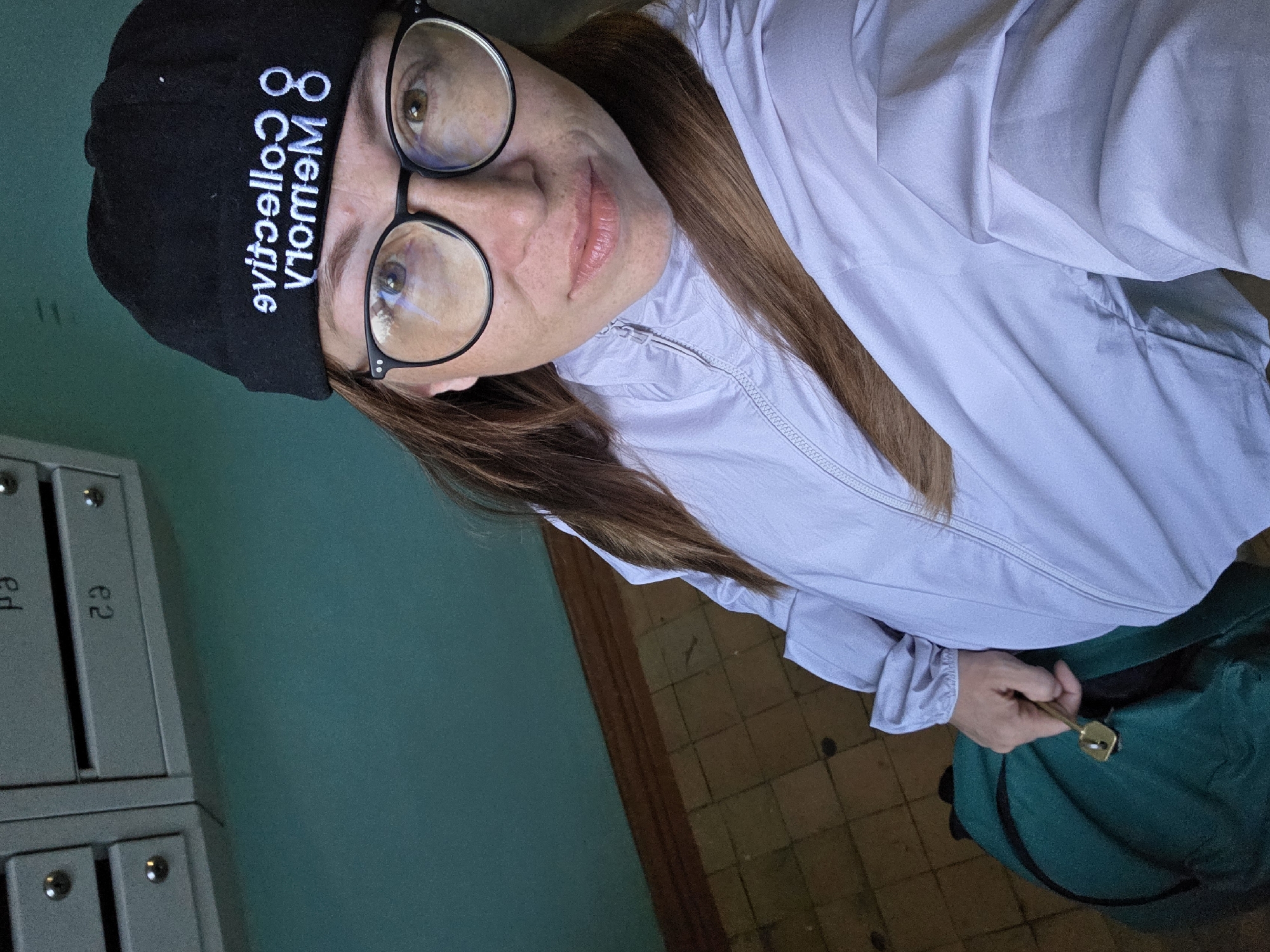 A woman, wearing a black beanie and glasses, stands in what appears to be an apartment building hallway, likely in Chaikovsky, Russia. The beanie has white text embroidered on it. She is wearing a light purple jacket and holding a green bag with a key. The background shows a section of mailboxes with numbers, and a turquoise-colored wall. The floor is tiled.