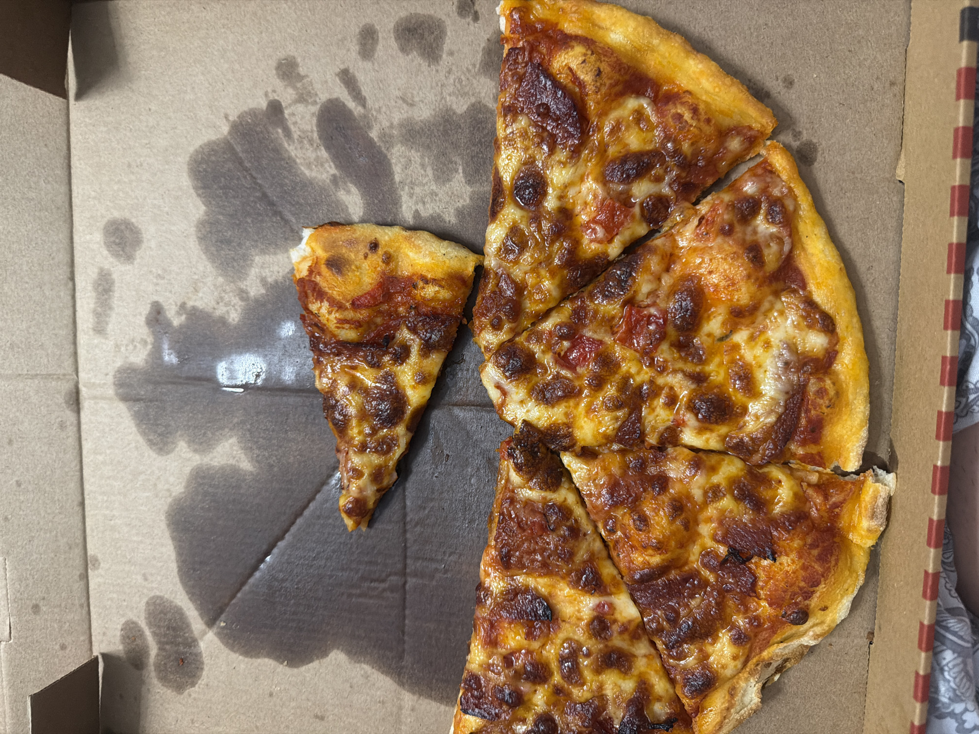 The image shows a pizza box with several slices of pizza inside. The pizza is a cheese pizza with a red sauce. There are some grease stains on the cardboard box. The image is taken from a top-down perspective. The pizza appears to have been partially eaten, with some slices missing. The pizza box is a standard cardboard pizza box. There are no people visible in the image. The environment appears to be indoors, possibly in a home or restaurant. The location is Ruse, Bulgaria.