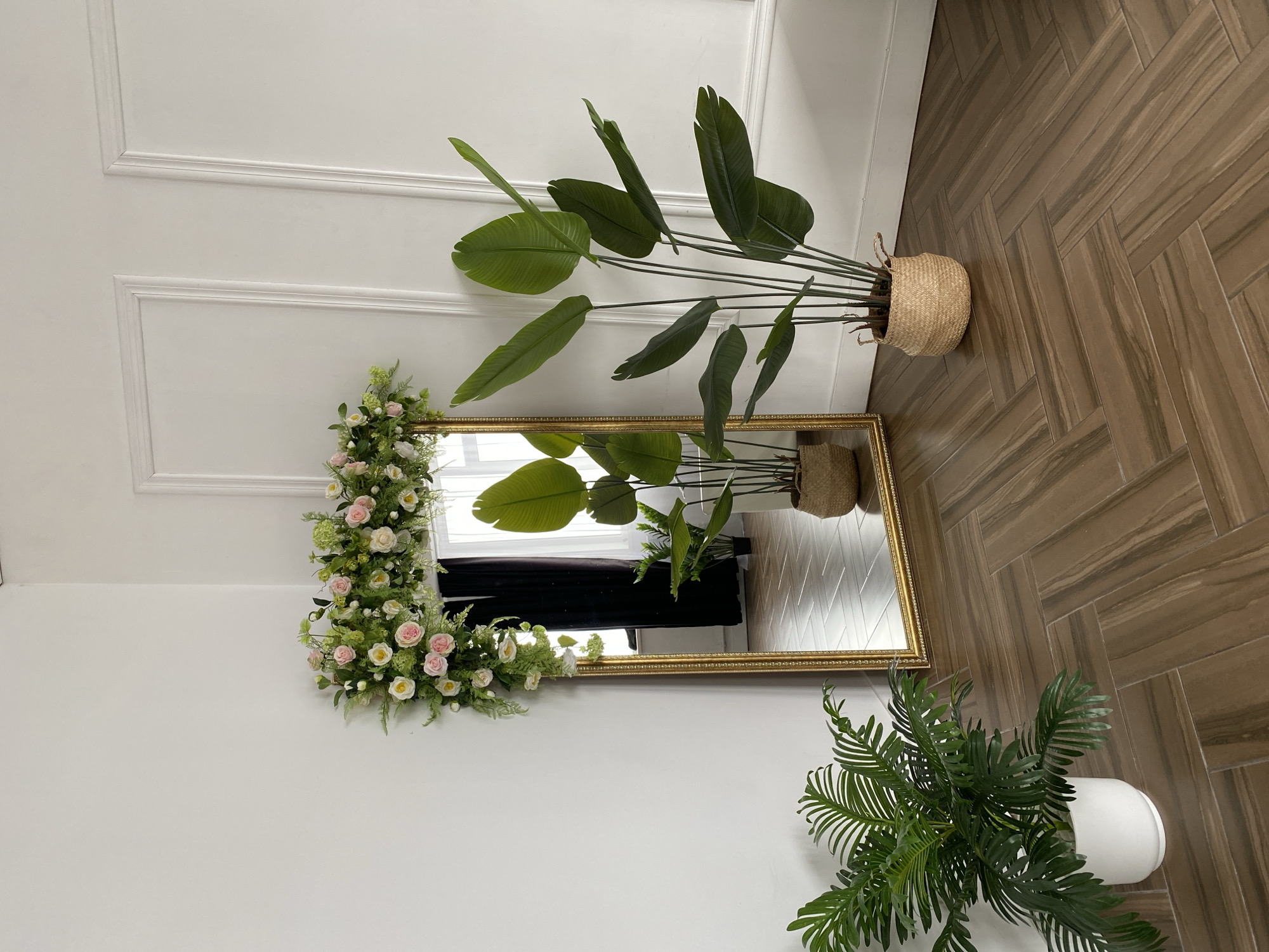 The image depicts an indoor setting with a focus on decorative elements. A gold-framed mirror adorned with flowers is positioned on a white wall with raised rectangular paneling. The mirror reflects a window covered by black fabric. To the upper right of the mirror, a tall plant in a woven basket stands on a herringbone-patterned wooden floor. A fern in a white pot sits in the lower right corner. The scene is brightly lit. There are no people visible in the image. The location of Chaikovsky, Russia, is not explicitly evident in the image.