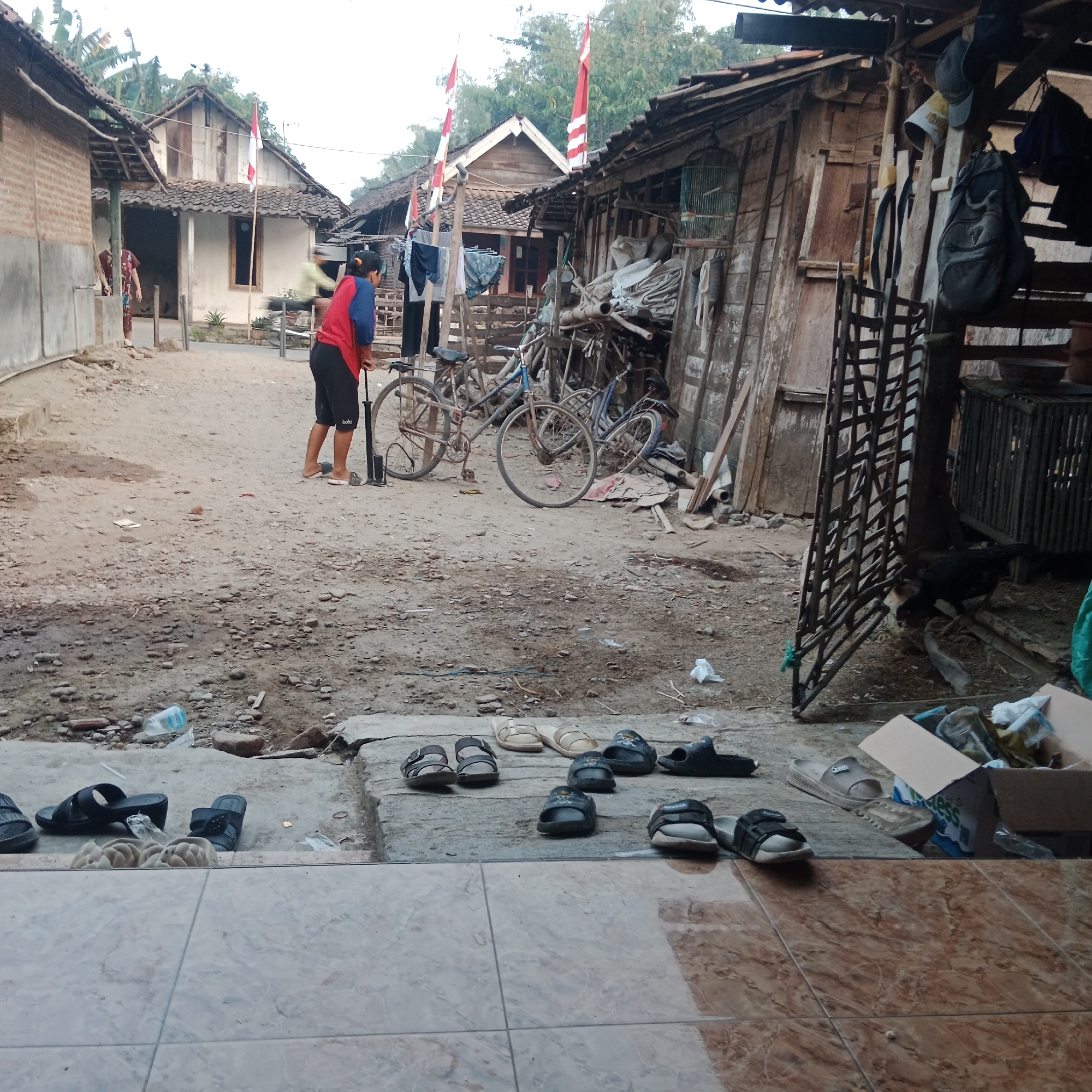 In the village of Ngringin, Indonesia, a scene unfolds on a dirt road in front of rural dwellings. A person in a red shirt and black shorts is seen using a bicycle pump, attending to one of the several old bicycles scattered around. Indonesian flags, red and white, are visible, suggesting a national holiday or celebration. Clothing hangs on a line, drying in the open air. Several pairs of sandals and shoes are arranged on the ground, indicating a residential area. The environment appears modest, with weathered wooden structures and a birdcage adding to the rustic ambiance. The overall impression is one of everyday life in a rural Indonesian community.