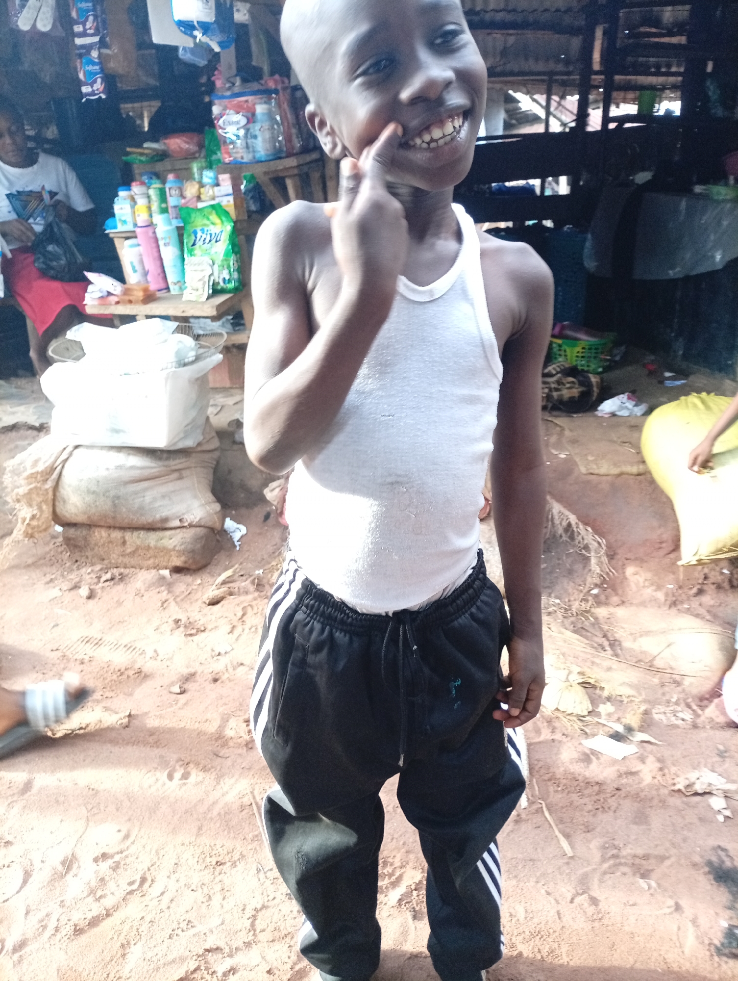 This is a full shot of a young boy, appearing to be between 8-10 years old, standing in what seems to be a market environment. The boy is wearing a white tank top and black track pants with white stripes on the sides. He has a very wide, toothy grin and is pointing to his face with his right index finger.
Behind the boy, the setting appears to be a market stall with various goods displayed on shelves. Items include bottles of liquids, bags of what might be snacks or other consumer products, and some bags of produce or grains. In the background, other people are visible, one woman in a white shirt and red skirt, and another person partially obscured. The ground is dirt and sand, indicating an outdoor or open-air setting.
The overall impression is a candid shot of a child enjoying themselves amidst the hustle and bustle of a local market in Nkpor, Nigeria. The lighting suggests it is daytime. The image has a slightly grainy quality, which is common in low-light or older digital photography.