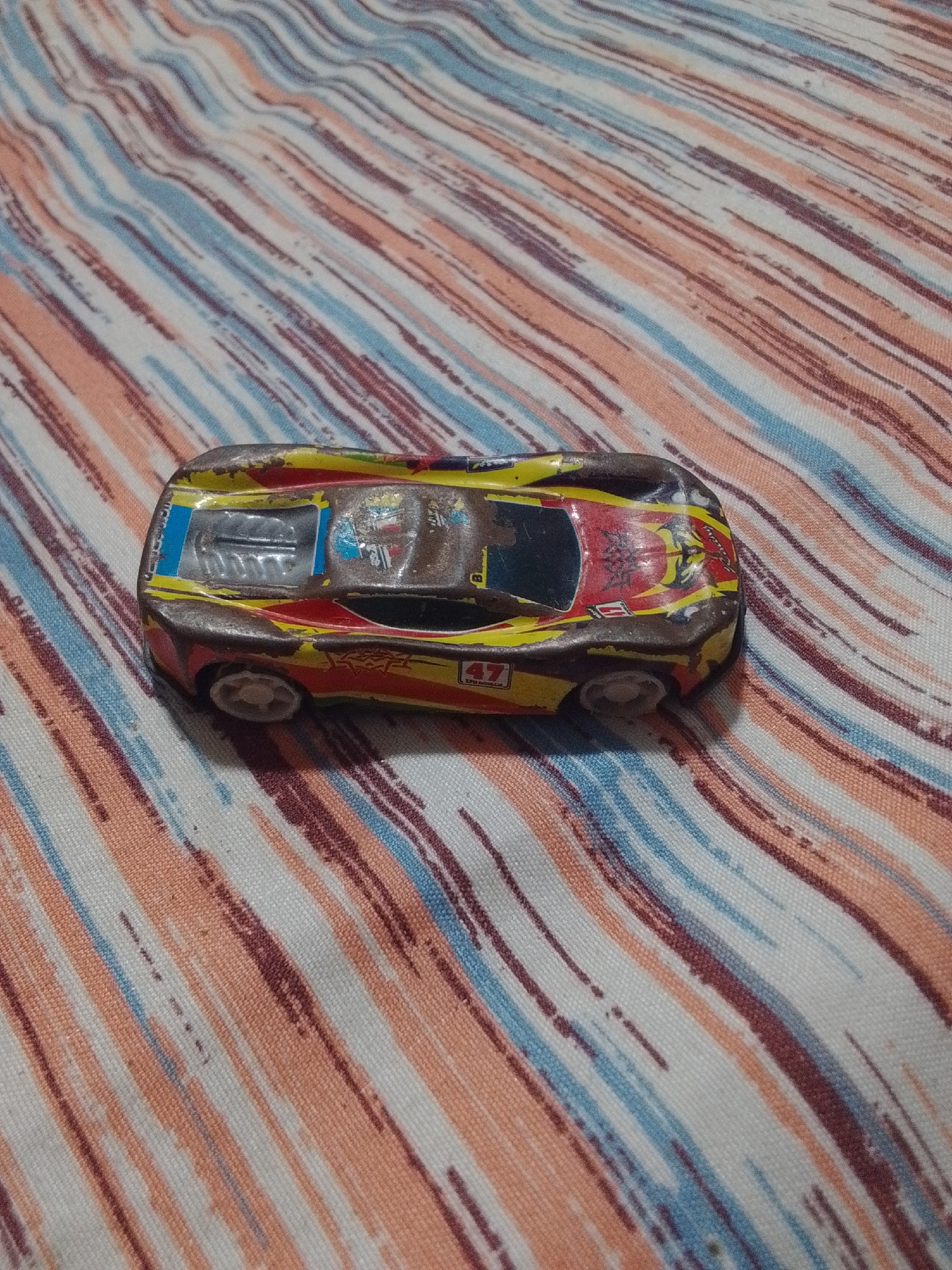 The image shows a close-up, eye-level shot of a toy race car. The car is a low-profile, futuristic design with a predominantly brown body. It features red and yellow racing stripes and decals, including the number "47" in a white box on the side. The car appears to be made of metal, possibly die-cast, and shows signs of wear, with some paint chipped away, revealing the underlying metal. The car has four white wheels, though the rear wheels are partially obscured by the car's body.
The car is resting on a striped fabric, which appears to be a tablecloth or bedsheet. The fabric has a pattern of irregular, diagonal stripes in shades of peach, light blue, and dark red/brown, set against a white background. The lighting is somewhat dim, with a soft, diffused light source coming from the right, casting subtle shadows. There are no people visible in the image. The setting appears to be indoors, on a flat surface. The overall mood is a simple, still life of a toy. No specific time of day, weather, or strong emotional cues are evident. There is text visible on the car, including "47" and what appears to be "SUPER RACING CAR" below it. Other small text and logos are present but illegible due to the resolution and wear on the toy. The context provided indicates the location as La Tagua, Venezuela, but this information cannot be visually inferred from the image itself.