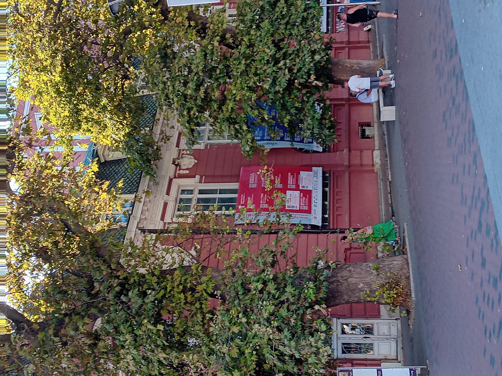 The image shows a street scene in Varna, Bulgaria. A large, leafy tree stands in the foreground, partially obscuring a red building with white trim and windows. There are several posters or banners hanging on the building. A woman in a green dress is walking past the tree. Further down the street, two men are sitting on a bench, and another man is walking by. The street is paved with cobblestones, and there are more trees and buildings in the background. The sky is visible at the top of the image.