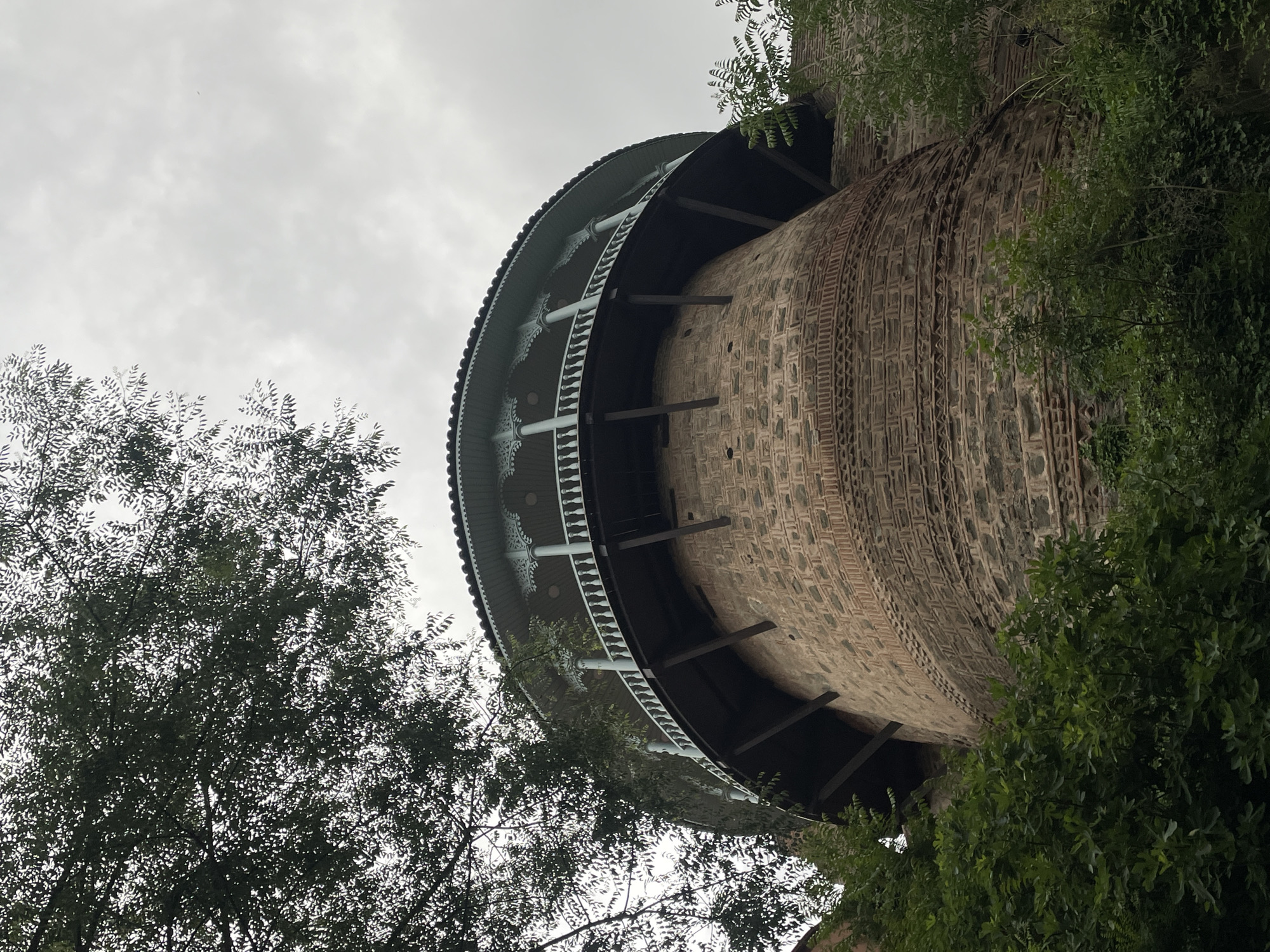 The image shows a low-angle view of the Narikala Fortress in Tbilisi, Georgia. The fortress tower is made of patterned brick and stone. A green and white circular viewing platform is partially visible at the top of the tower, supported by dark beams. Trees and foliage surround the structure. The sky is overcast.