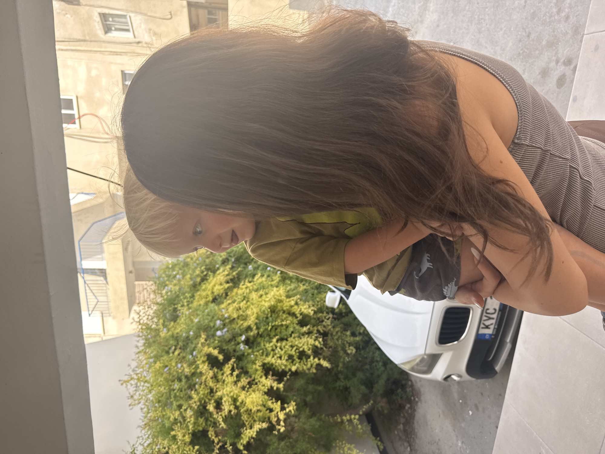 An over-the-shoulder shot shows a woman holding a young child. The child, with blonde hair and blue eyes, is looking to the left, with their mouth slightly open. The woman has long, dark brown hair and is wearing a striped tank top. In the background, a white car is parked in front of a building with balconies. Lush green foliage is visible to the left of the car. The scene appears to be outdoors, likely on a balcony or patio, with a street or driveway visible below. The location is identified as the Agios Tychona Community, Cyprus.