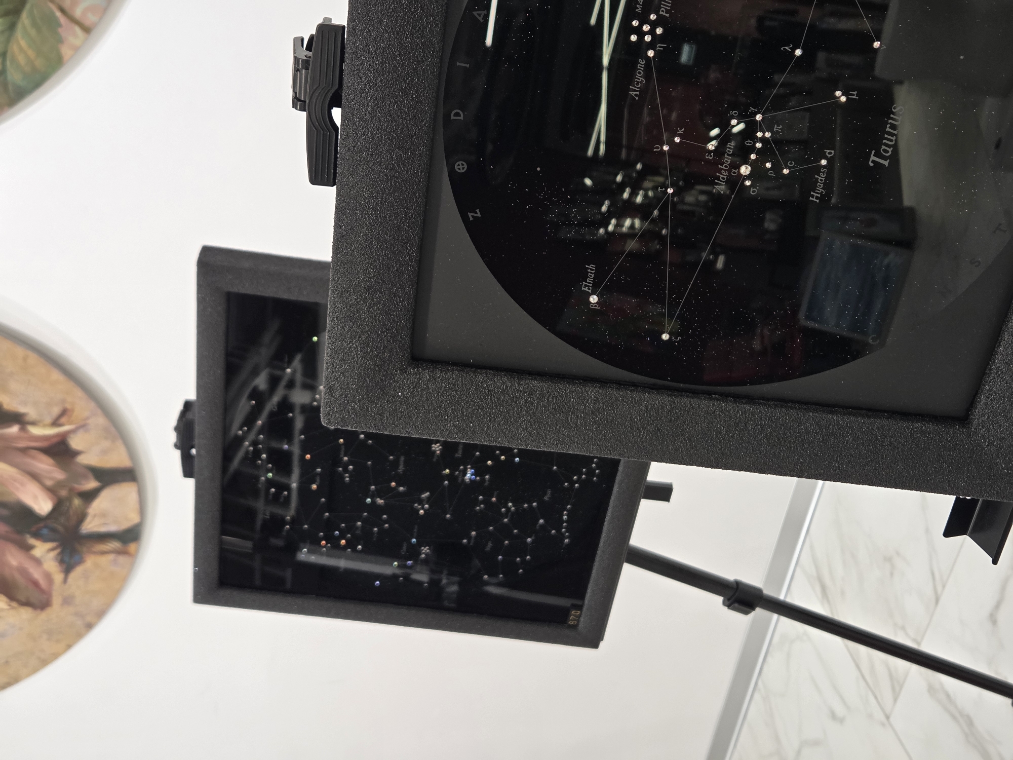 The image shows two electronic displays showing star constellations, likely in a museum or educational setting. The display in the foreground shows the constellation Taurus, with stars labeled "Aldebaran" and "Hyades." The display in the background shows a broader view of constellations, including one labeled "Leo." The displays are housed in black frames and are mounted on stands. The environment appears to be indoors, with a white wall and a patterned floor visible. The upper part of the image shows a portion of a circular painting of flowers. There are no people visible in the image. The scene does not provide any specific indicators to pinpoint the location to Minsk, Belarus.