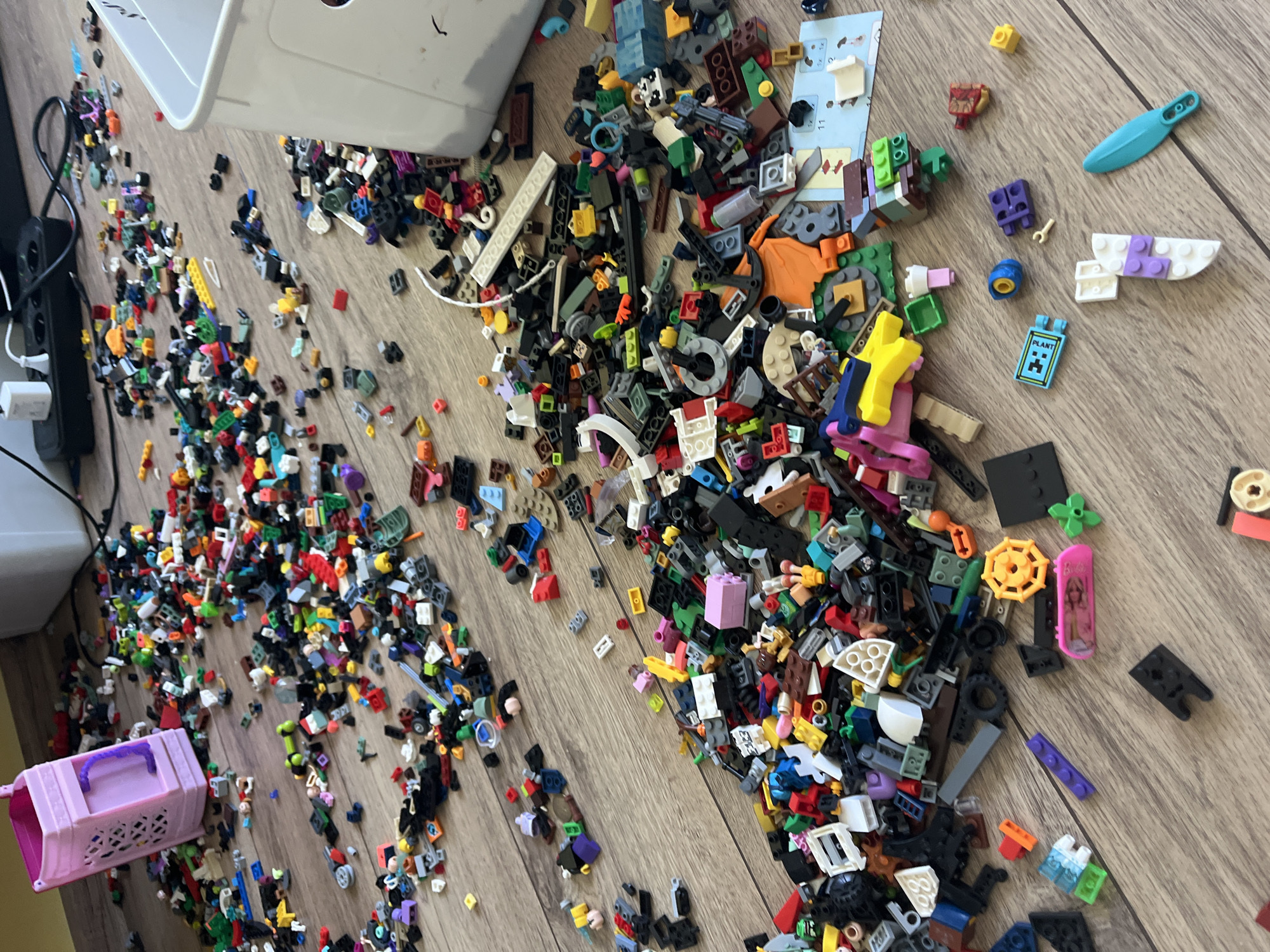 The scene depicts a large quantity of LEGO bricks scattered on a light brown wood-grain floor. The bricks are of various colors, sizes, and shapes. A partially visible white plastic bin and a pink toy storage container with a purple handle are also present. A power strip with plugs is visible along the left edge. The location is a room with wood flooring.
