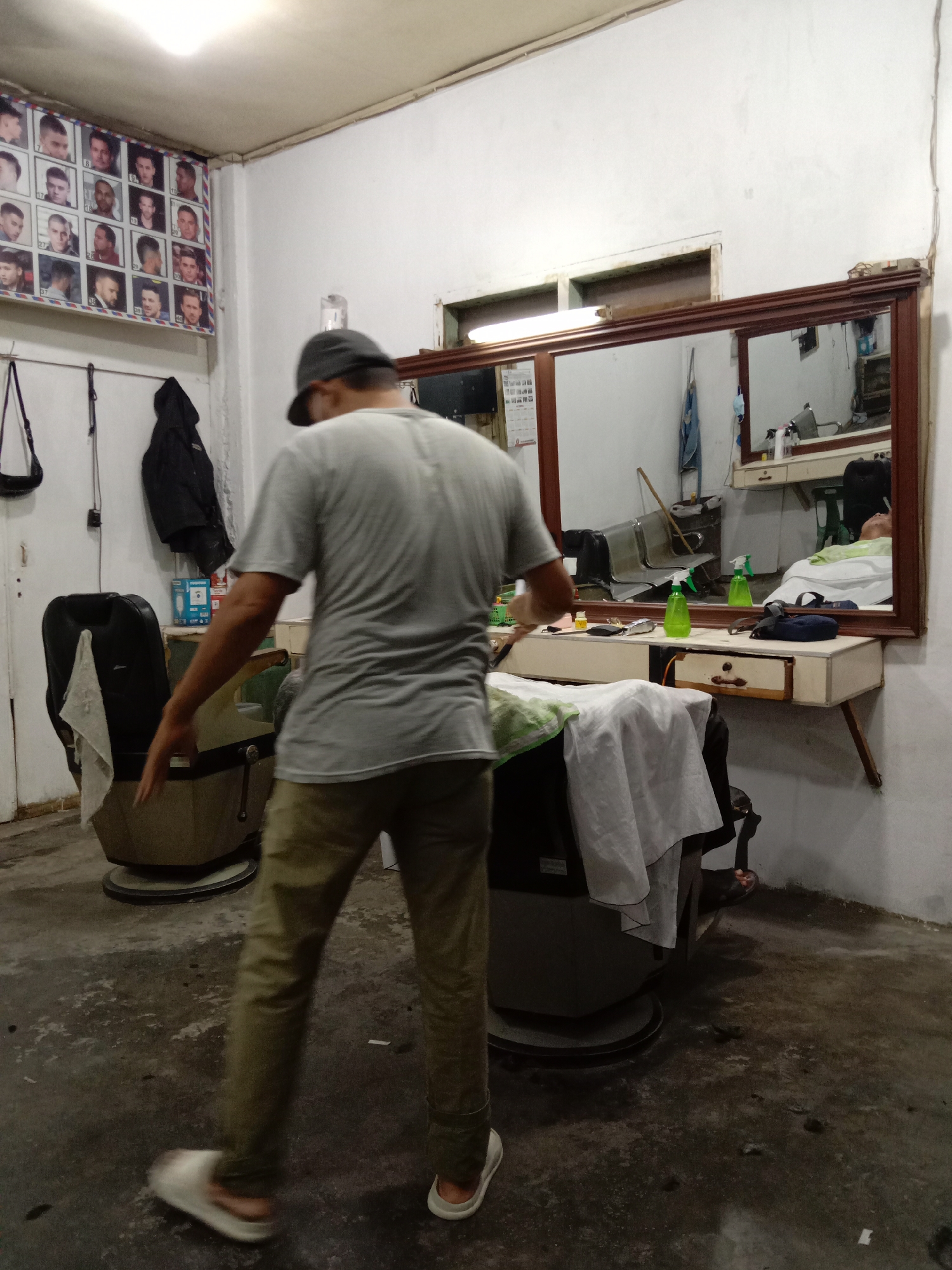 The image shows the interior of a barber shop in Lhokseumawe, Indonesia. A man wearing a gray t-shirt, khaki pants, and a baseball cap is walking away from the camera. Two barber chairs are visible, one with a white cloth draped over it. A counter with a large mirror hangs on the wall. Various grooming supplies, including green spray bottles, are on the counter. A poster displaying different hairstyles is on the wall. Clothes hang near the corner of the room. The floor appears to be dark and somewhat unclean, littered with hair. A customer is visible in the mirror in a chair, lying down, presumably for a treatment.