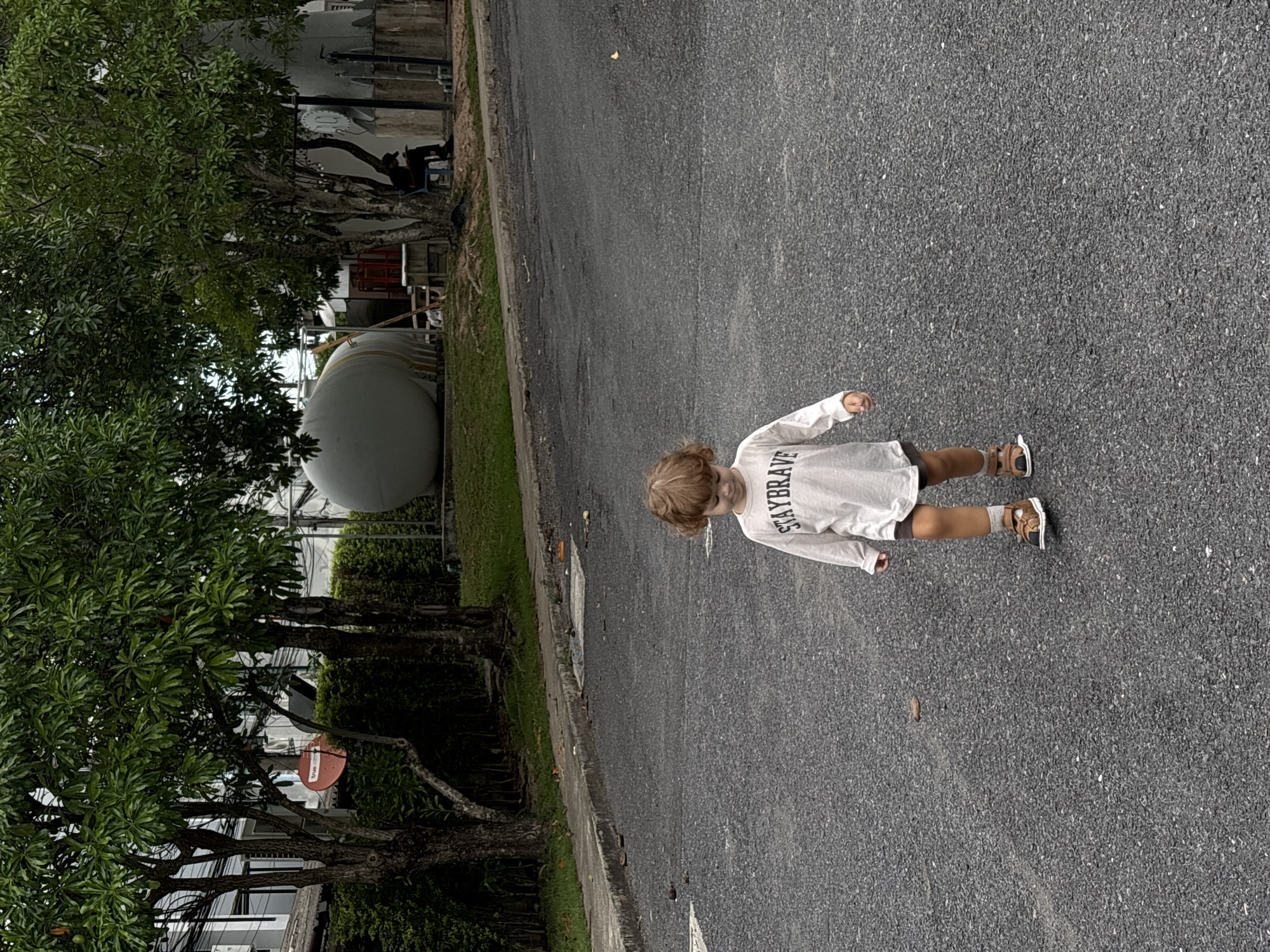 A full shot depicts a small child walking on a paved road. The child has blonde hair and is wearing a white, long-sleeved shirt with the words "STAYBRAVE" printed on it. The child is also wearing brown shorts and sandals with light-colored socks. The road is grey and appears to be asphalt. To the left of the road, trees and green vegetation are visible. Beyond the trees, a large, spherical, grey object is present. A red satellite dish is mounted on a tree. The location is consistent with a street scene in Choeng Thale, Thailand.
