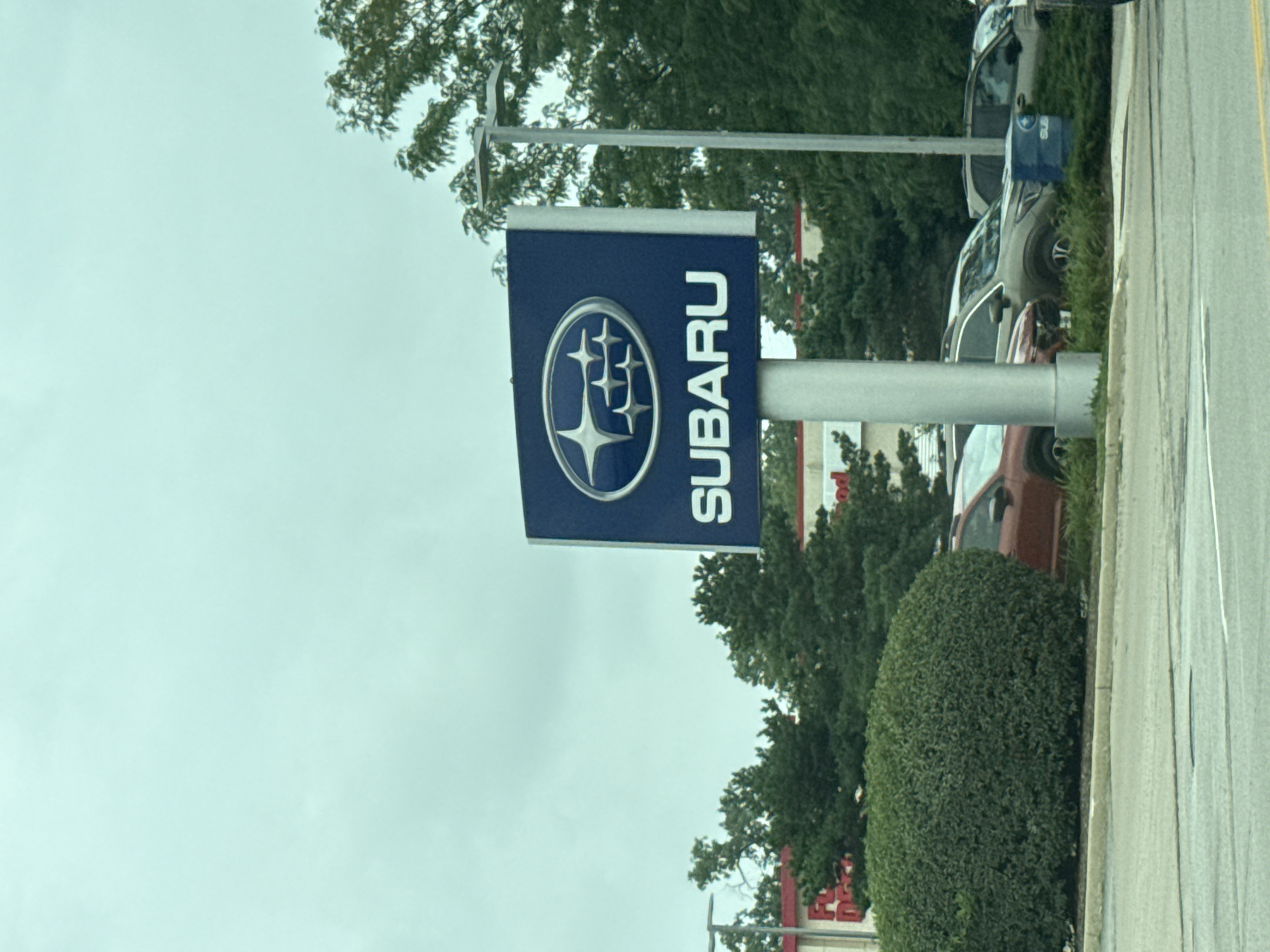 The image shows a Subaru dealership sign with the Subaru logo and the word "SUBARU" in white lettering on a blue background. The sign is mounted on a silver pole. In the background, there are several Subaru vehicles parked, including a red Subaru Forester, a tan Subaru Outback, and a dark gray Subaru Forester. There are also trees and bushes surrounding the dealership. The sky is overcast. The image was taken in Skokie, United States of America.